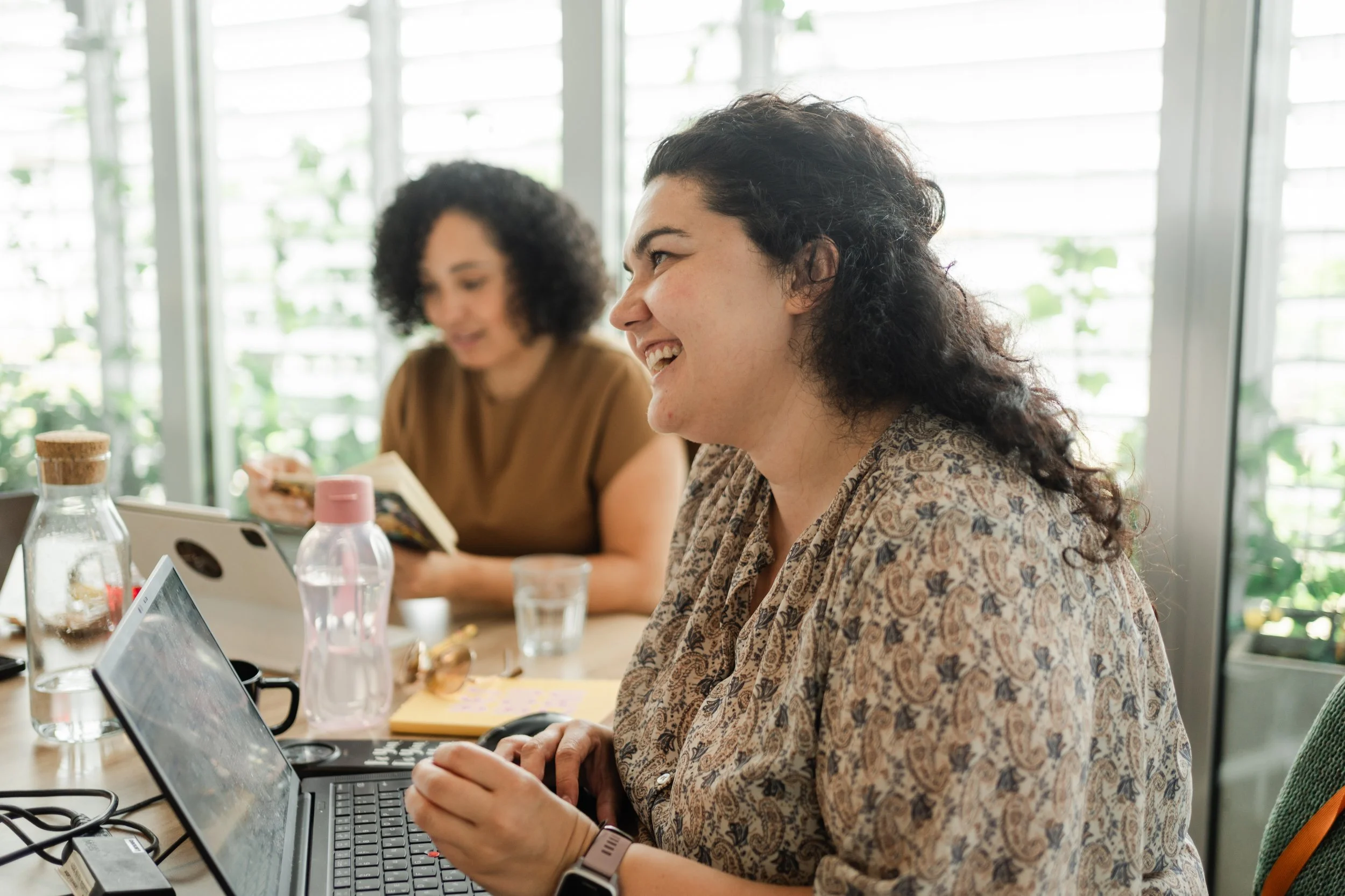 Two women with curly hair sitting at a table, smiling, with laptops, water bottles, and snacks in a bright room with large windows and greenery outside.