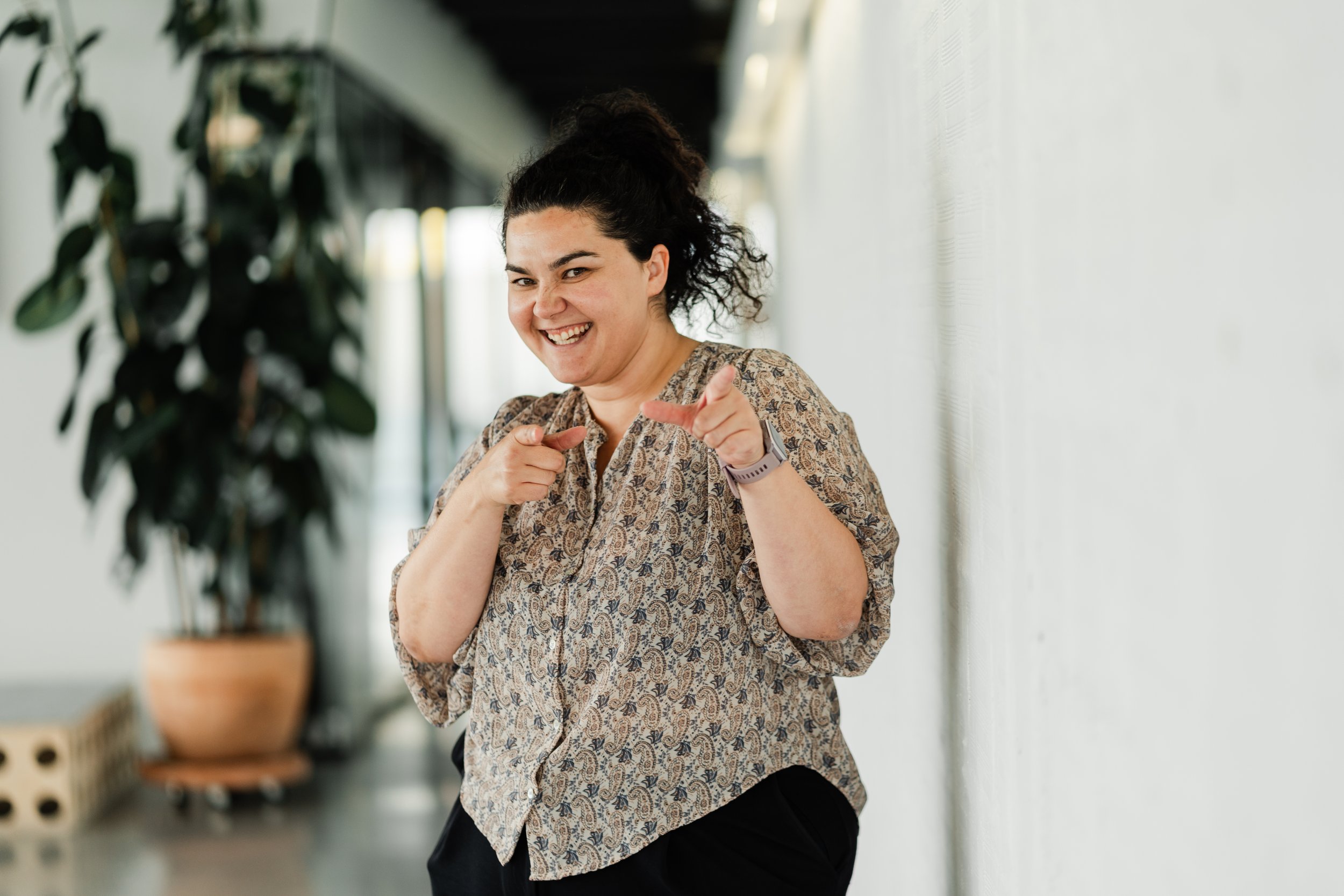 Smiling woman pointing with both hands indoors, near a white wall and a plant.
