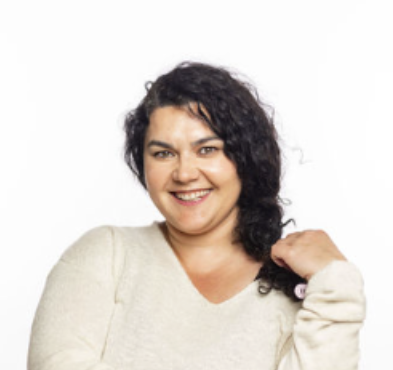 A woman with curly dark hair wearing a light-colored sweater, smiling and touching her shoulder against a plain white background.
