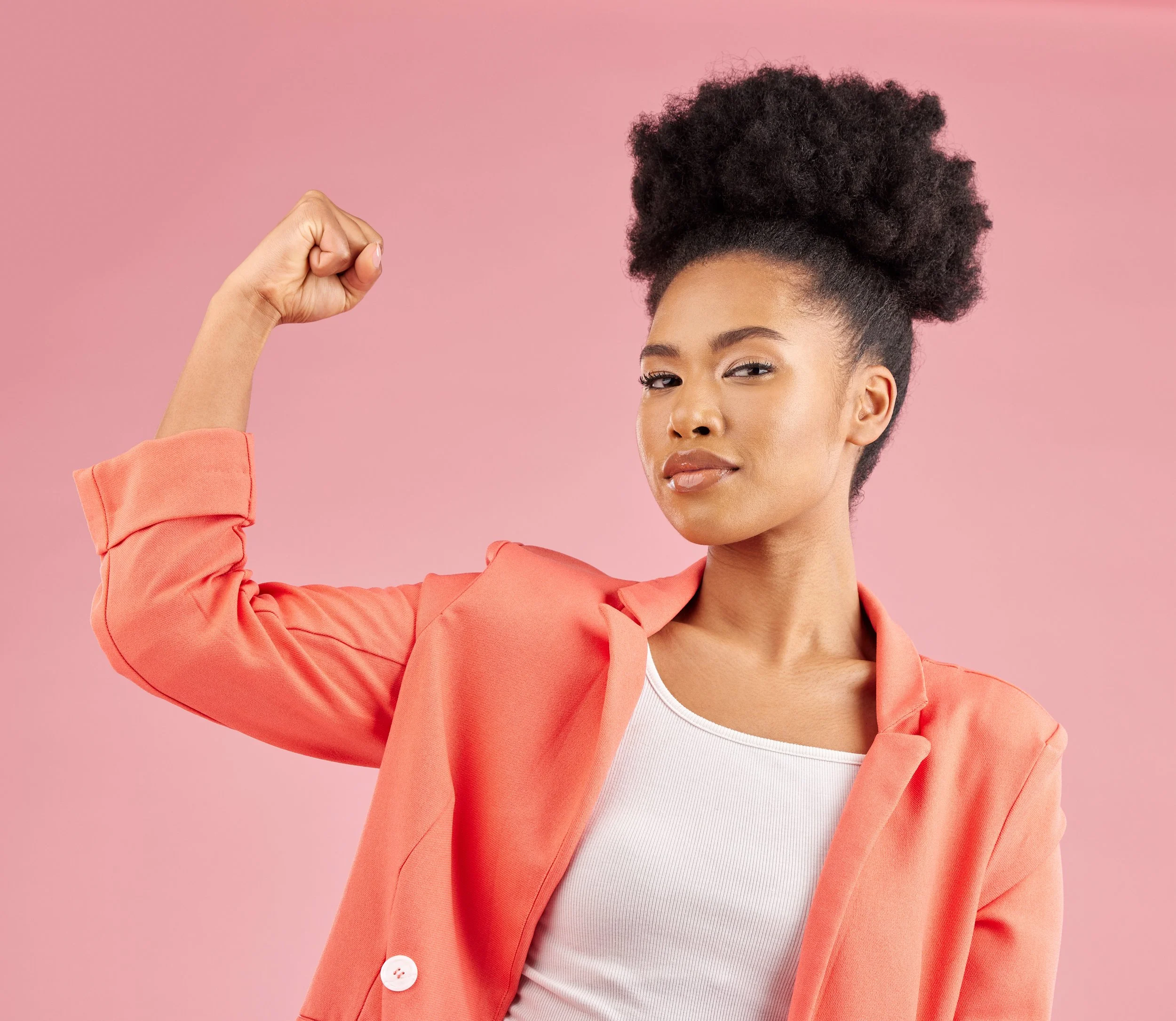 A woman flexing her arm with a clenched fist, wearing a coral blazer and white top, against a pink background.