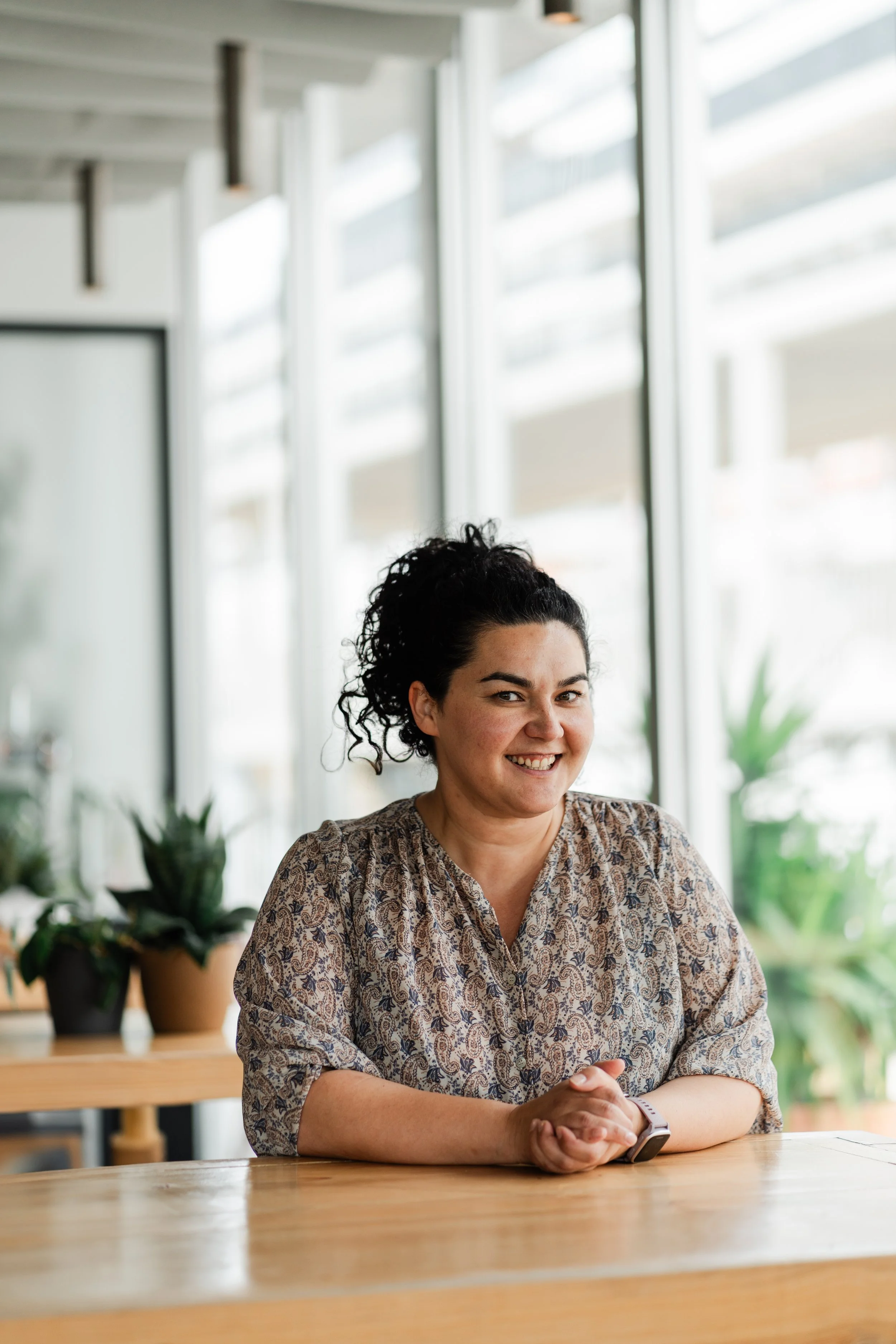 A woman with dark, curly hair in a ponytail, wearing a patterned blouse, sitting at a wooden table in a bright, modern room with large windows and potted plants, smiling and looking at the camera.