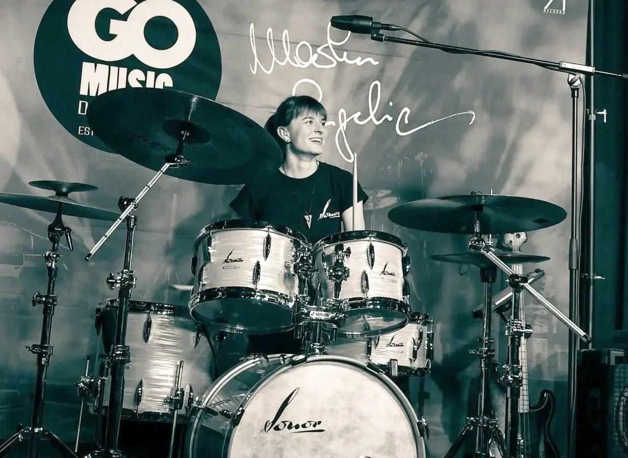 A woman sitting behind a drum set on a stage, smiling, with a go music banner and microphone.