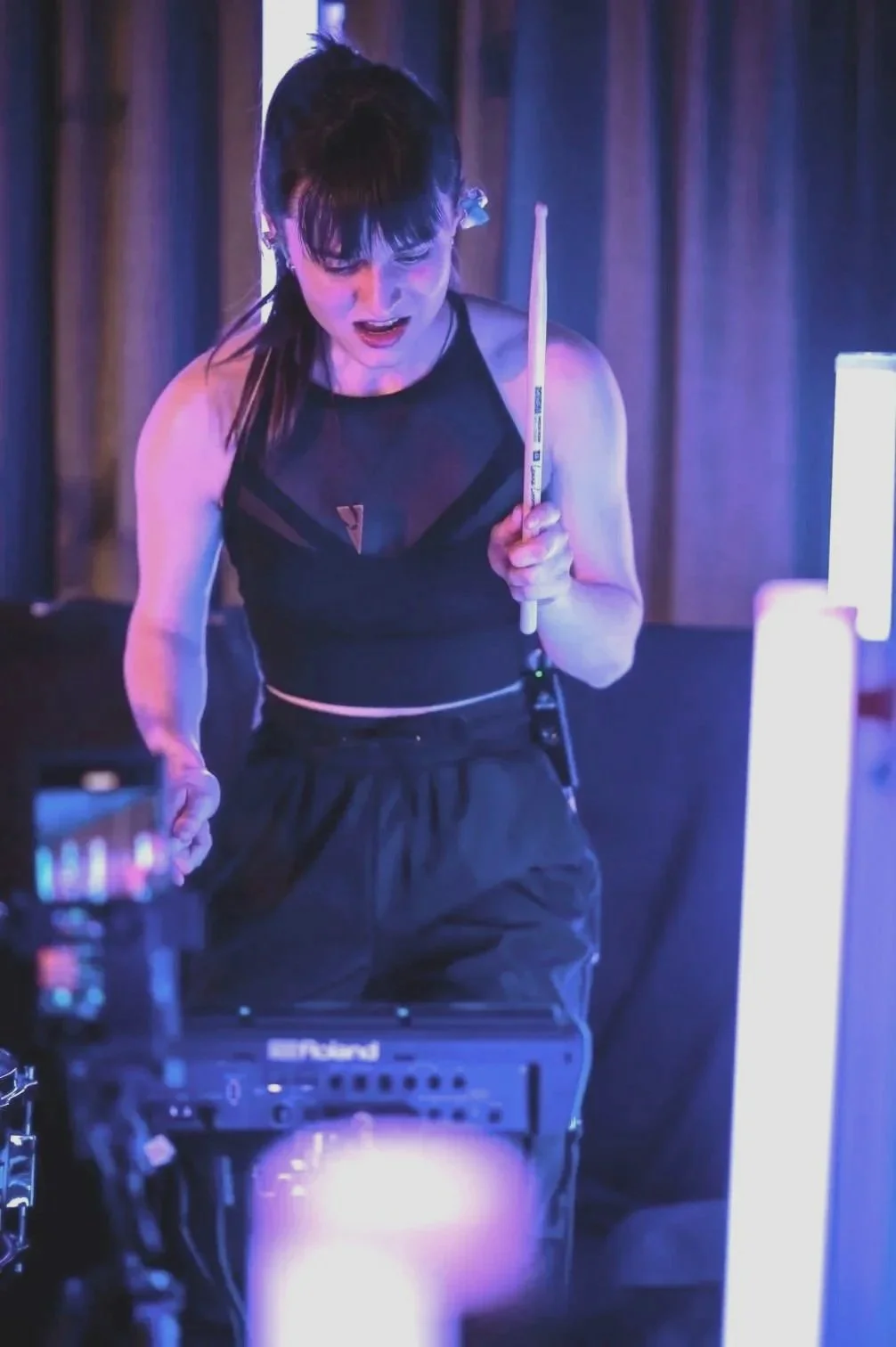 A woman playing pool with a cue stick in a dimly lit room, with neon lighting and a computer or sound equipment in front of her, wearing a black tank top and black pants.