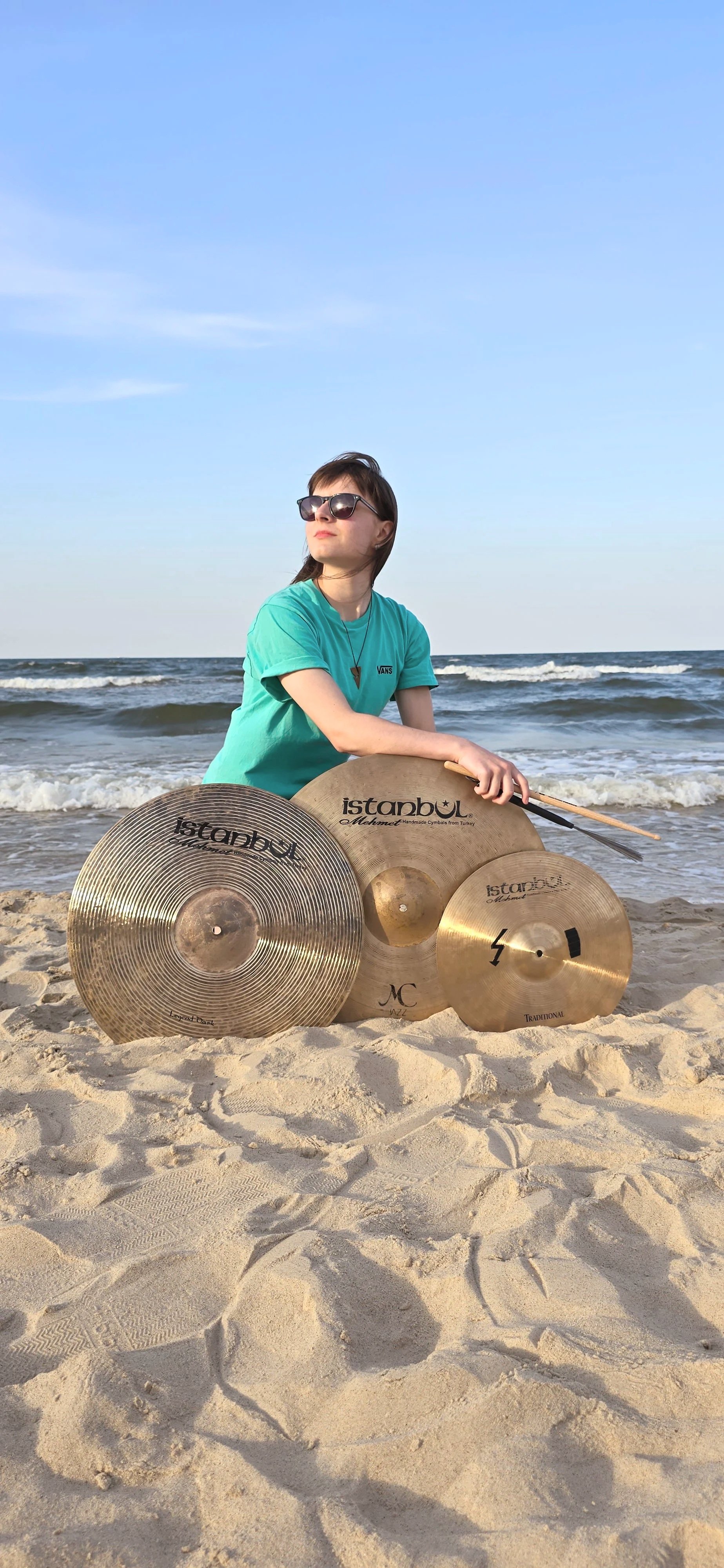 Woman sitting on the sandy beach holding drumsticks with three cymbals nearby, ocean waves in the background, wearing sunglasses and a teal T-shirt.