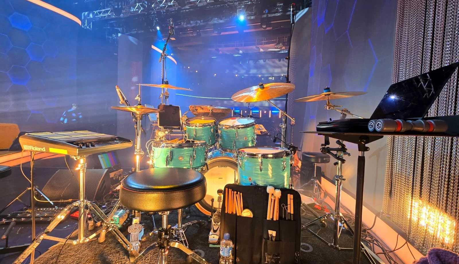 Music stage setup with drum set, electronic pads, laptop, microphone, and percussion instruments ready for a performance.