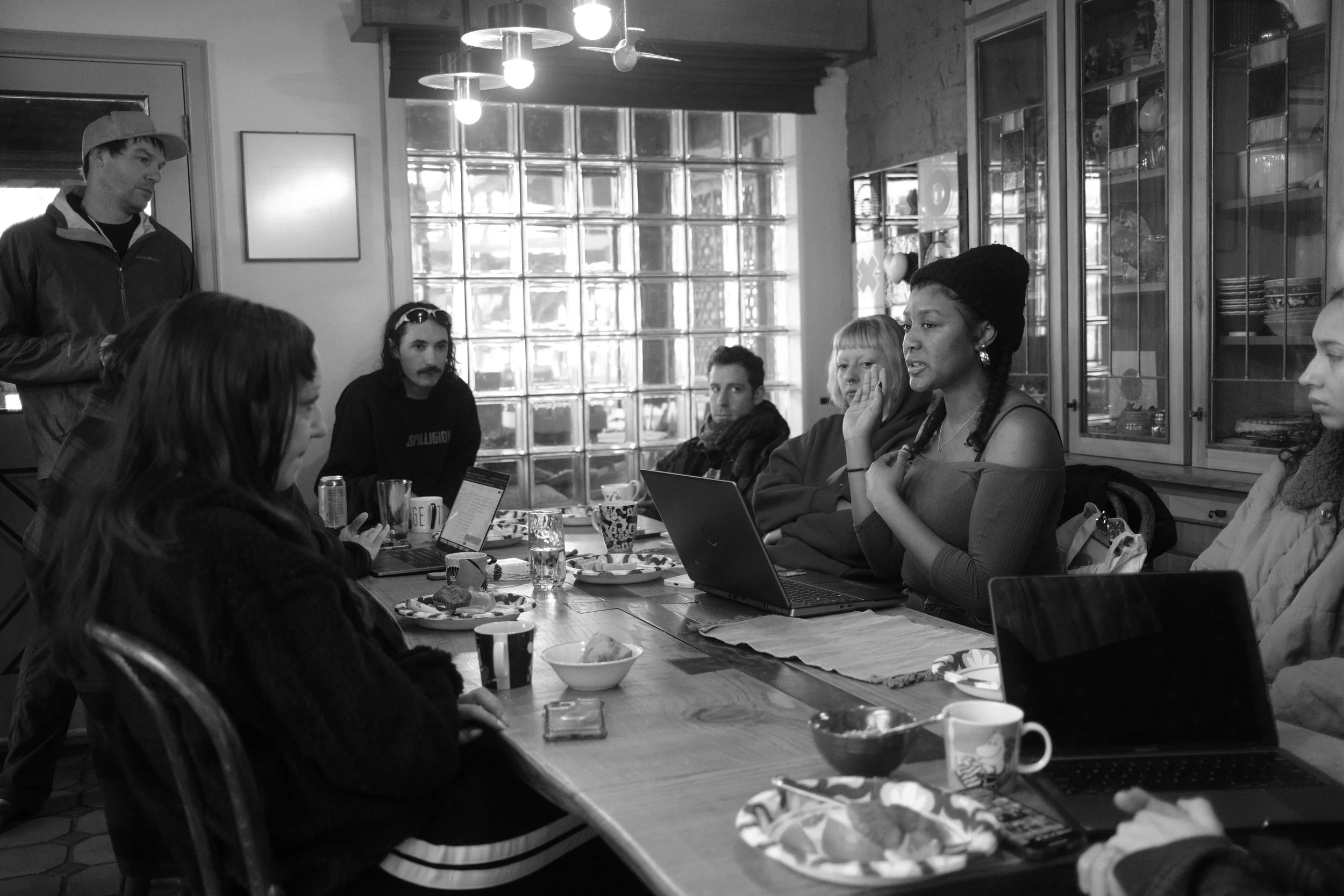 A group of people sitting around a table in a cozy cafe, engaged in conversation. Some are using laptops, and there are drinks and snacks on the table.