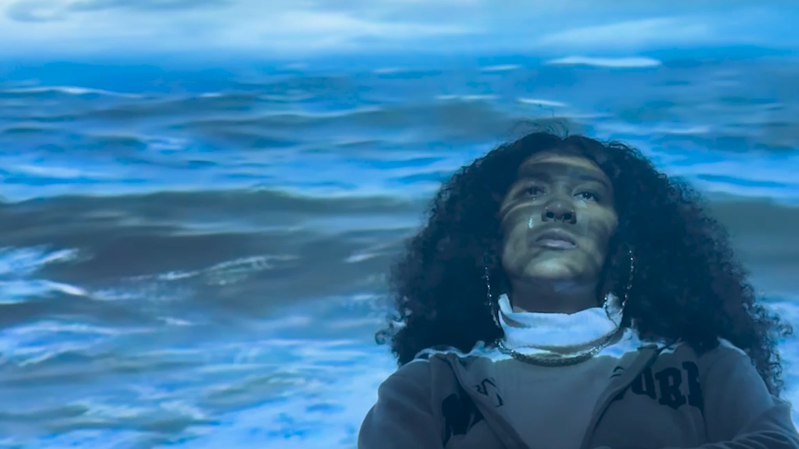 Person with curly hair wearing a face mask around the neck, standing in water near the ocean, looking upwards.