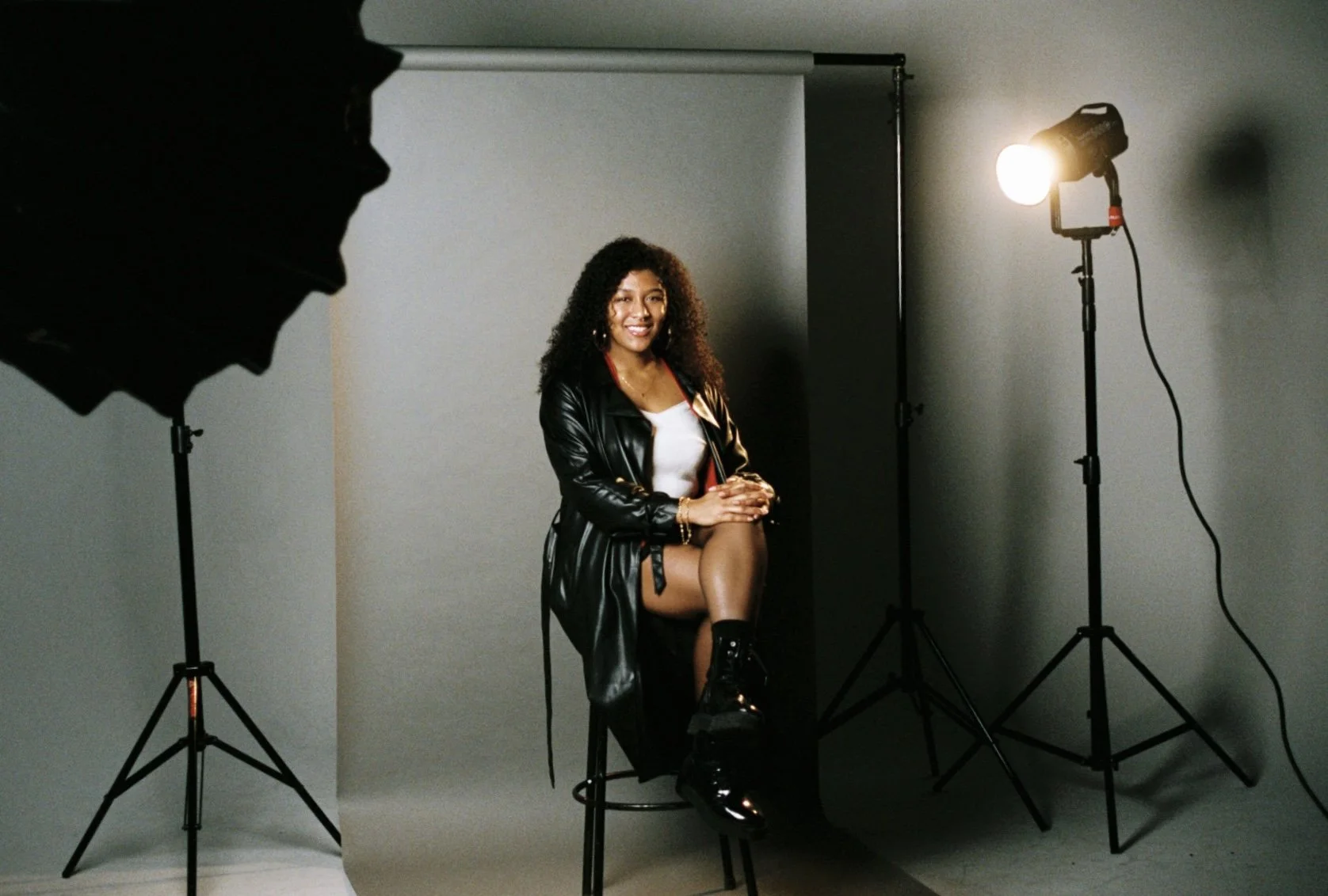 A woman sitting on a stool in a photography studio, illuminated by professional lighting, with a grey backdrop behind her.