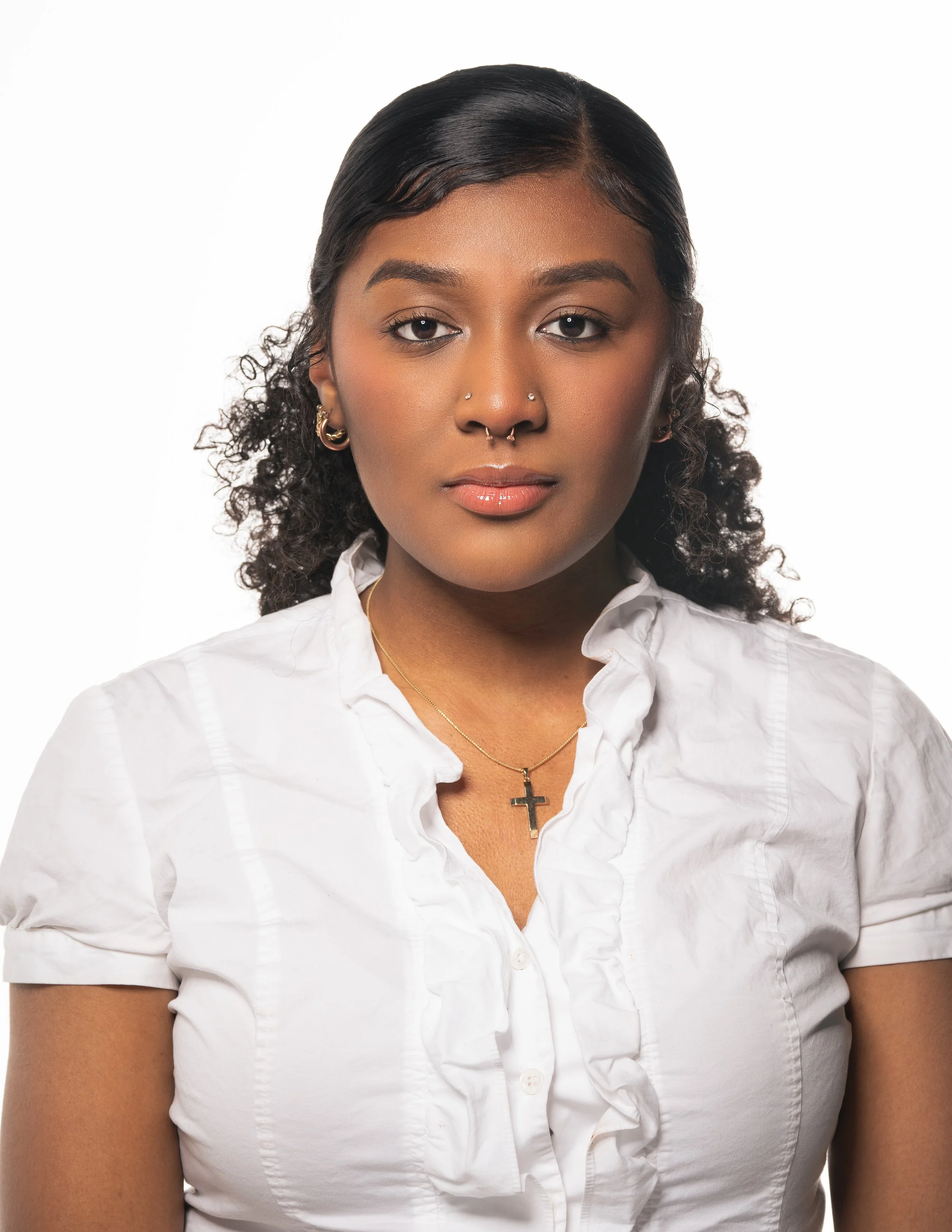 A woman with dark, wavy hair styled to the side, wearing a white collared shirt with ruffled detailing, gold hoop earrings, multiple facial piercings including a nose ring, septum ring, two nostril piercings, and a cross necklace, standing against a plain white background.