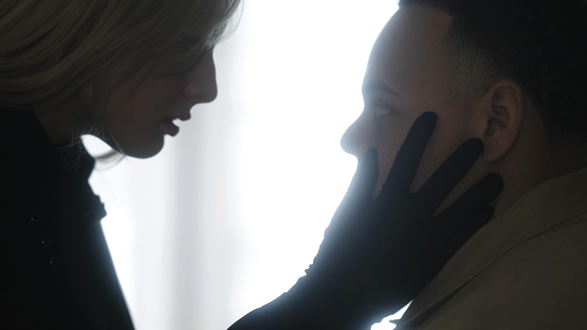 A person with blonde hair and a person with short, dark hair are close to each other, with the person with dark hair gently touching the face of the blonde-haired person, both silhouetted against a bright light background.