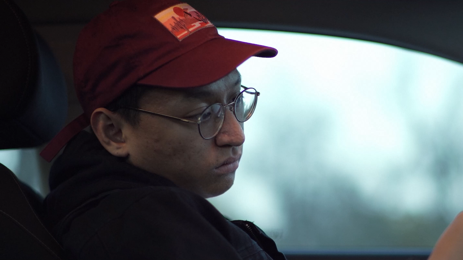 A young man with glasses and a red cap sitting in a vehicle, facing sideways with a blurred view outside the window.