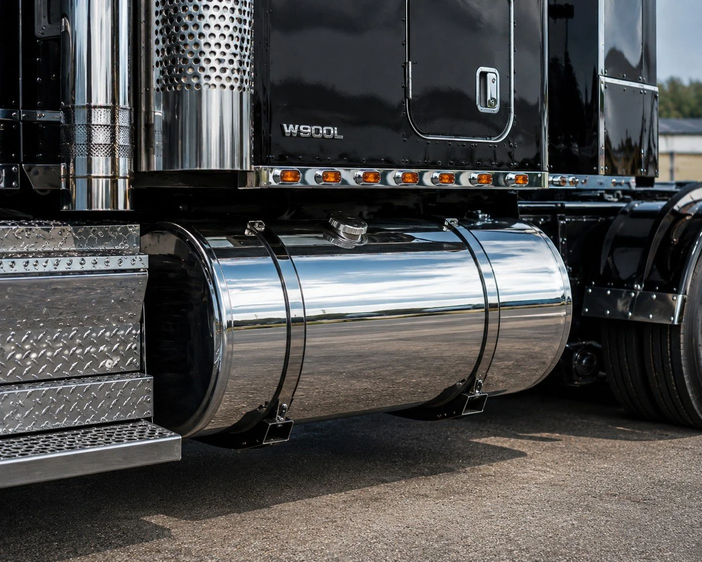 Close-up of a shiny, chrome fuel tank and part of a black semi-truck with chrome details, parked on asphalt with a house and trees in the background.