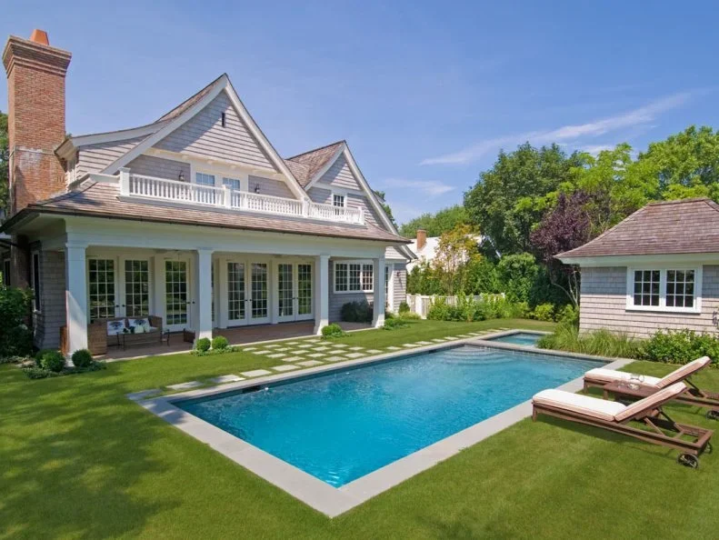 A backyard with a swimming pool in front of a large, two-story white house with a porch and balcony, surrounded by lush greenery and a small separate shed.
