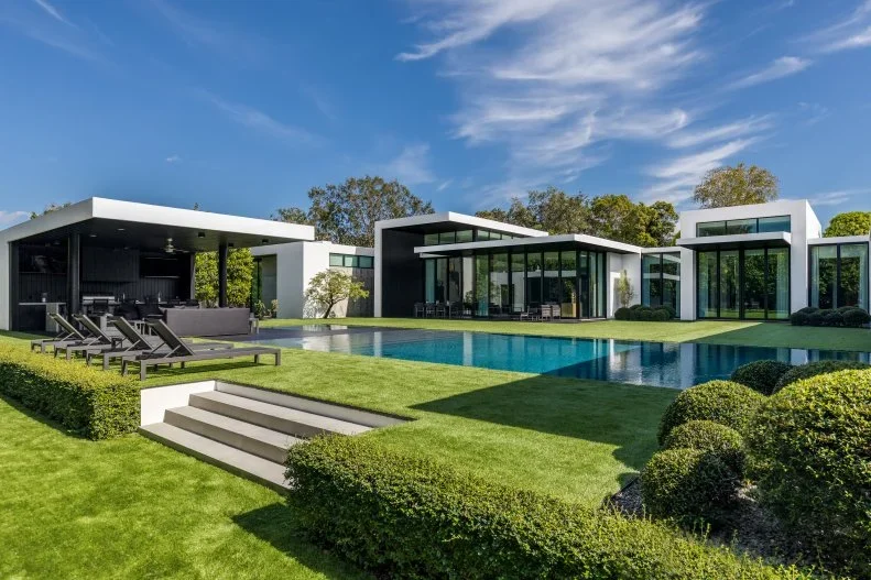 Modern luxury house with large swimming pool, outdoor lounge, and lush green lawn under blue sky.