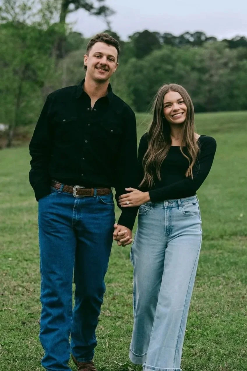 A young couple holding hands outdoors in a green park, smiling at the camera. The man is wearing a black shirt and jeans, and the woman is dressed in a black top and light-wash jeans.