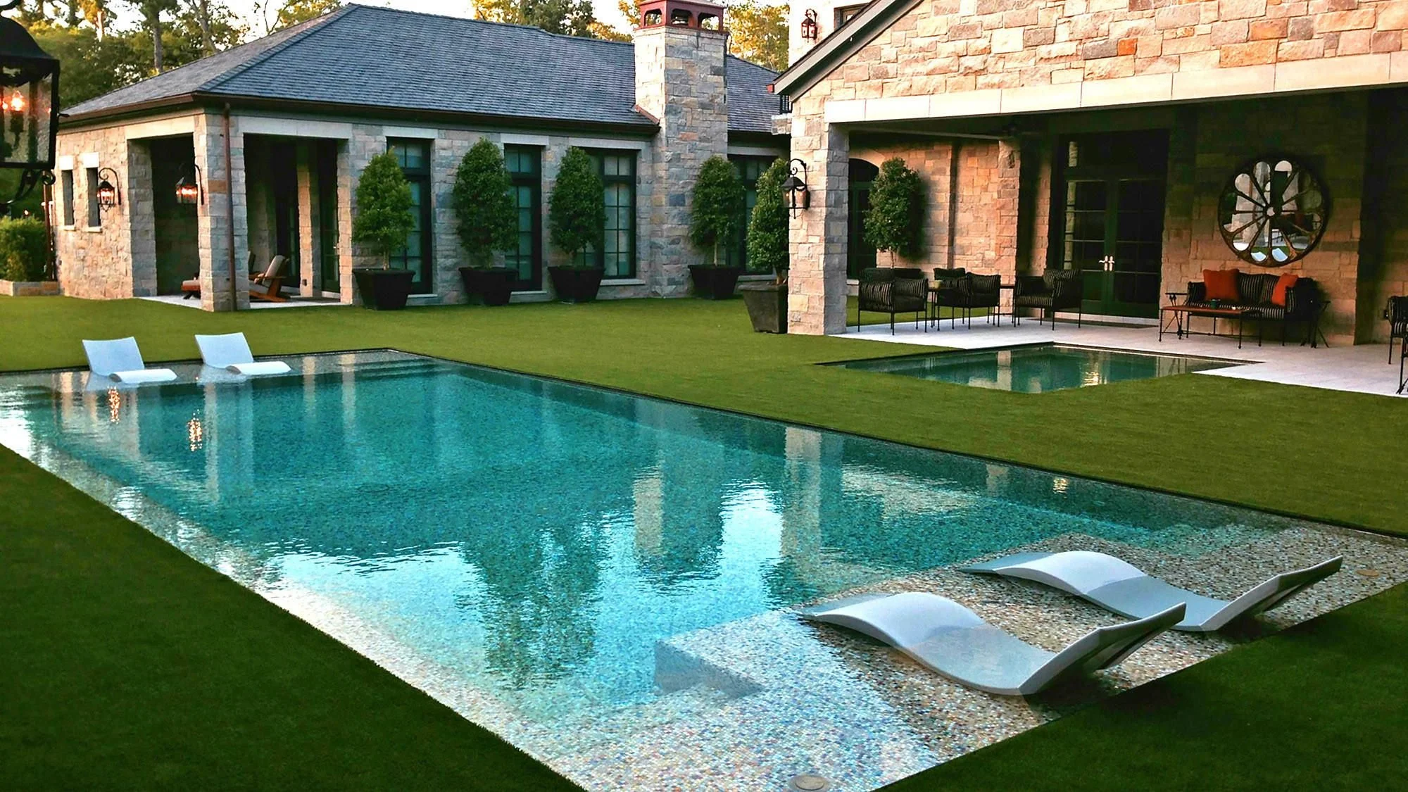 Backyard with a swimming pool, surrounded by a well-manicured grass lawn, outdoor seating, potted plants, and a stone house with large windows and covered patios.