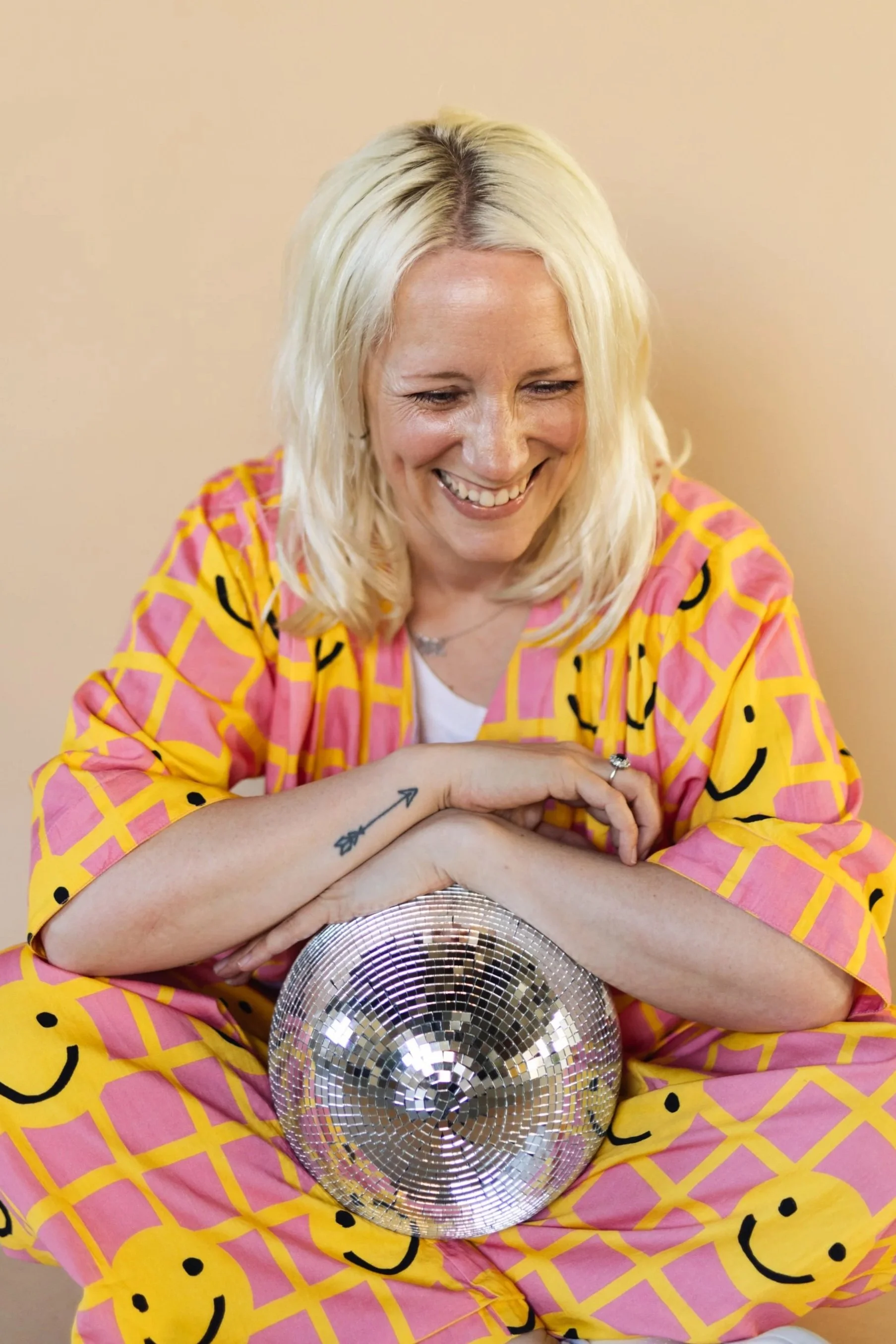 A blonde haired white woman sits cross legged, laughing at something while she leans on a disco ball in her lap. She is wearing a pink suit with yellow smiley faces on it, and has a tattoo of an arrow along the edge of her wrist.