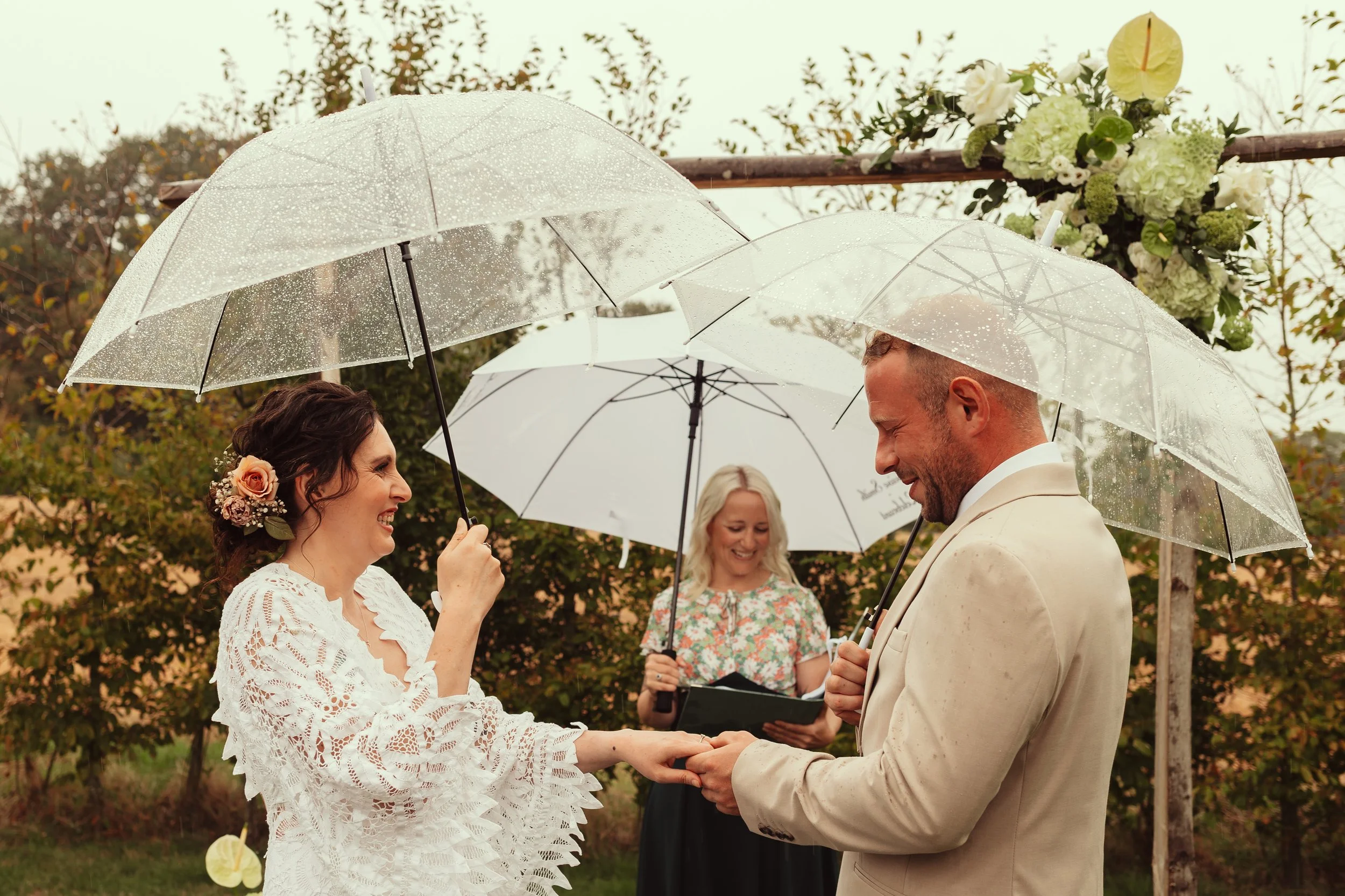 Two people exchanging rings with a celebrant in background, a white woman with dark hair in a vintage lace wedding dress and a fair haired white man in a light coloured suit.  All 3 are holding umbrellas.