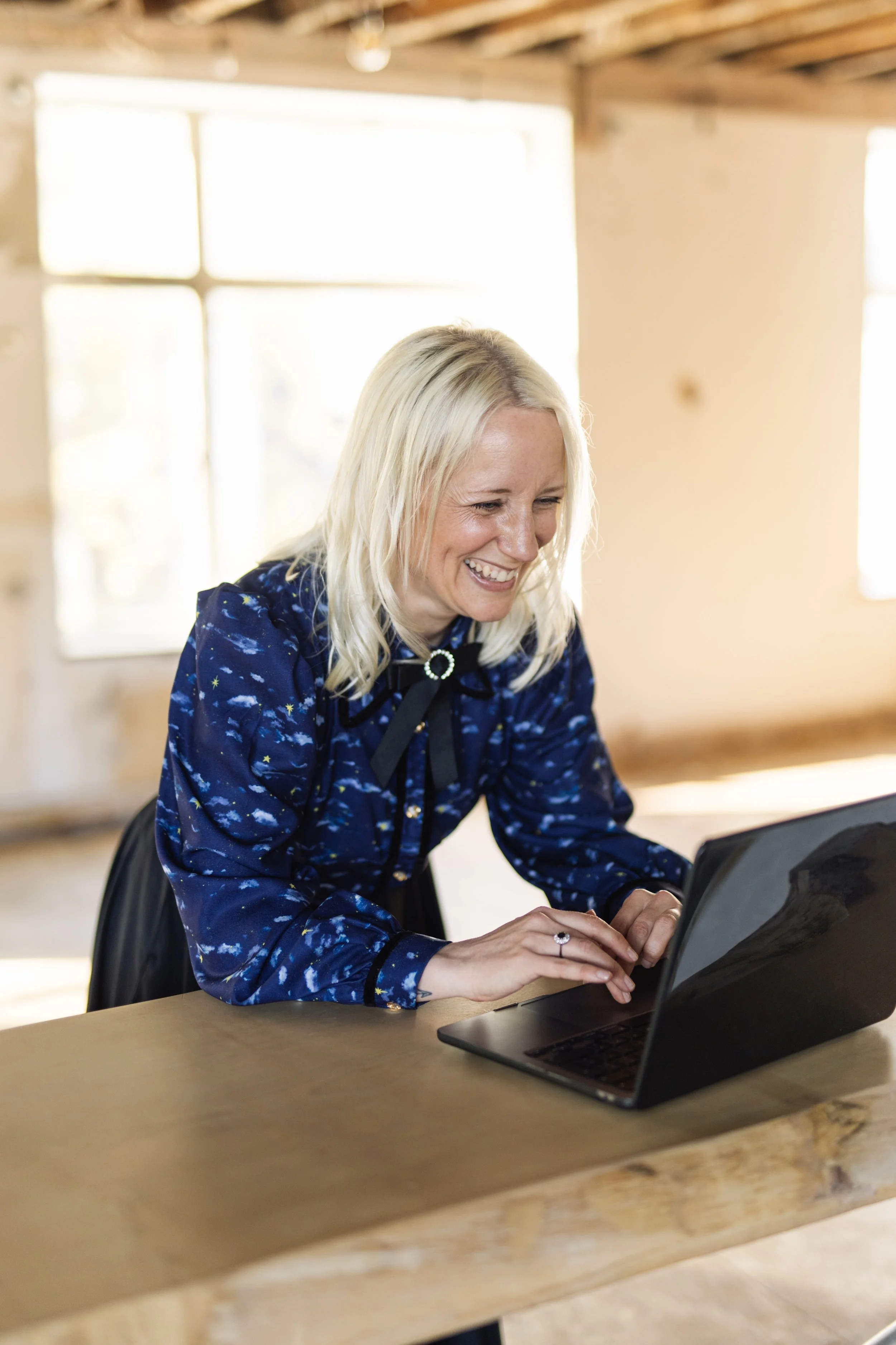 A blonde white woman in a dark blue blouse laughs as she looks at her laptop screen