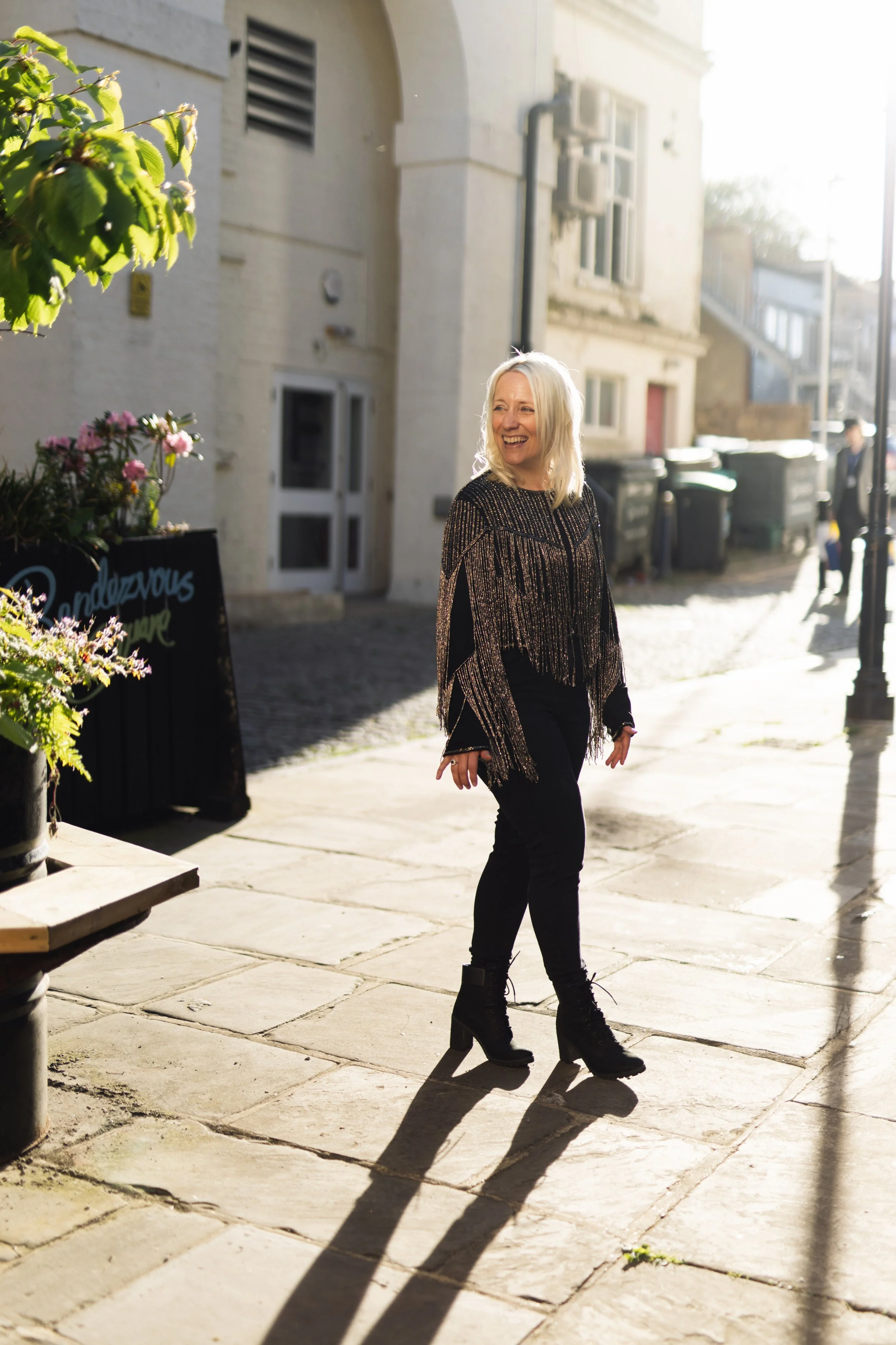 A white woman with blonde hair is wearing a black jacket with sparkly silver tassels on it, black skinny jeans and black boots with a chunky heel. She is in a sunny courtyard, and is walking off towards the right of the shot.
