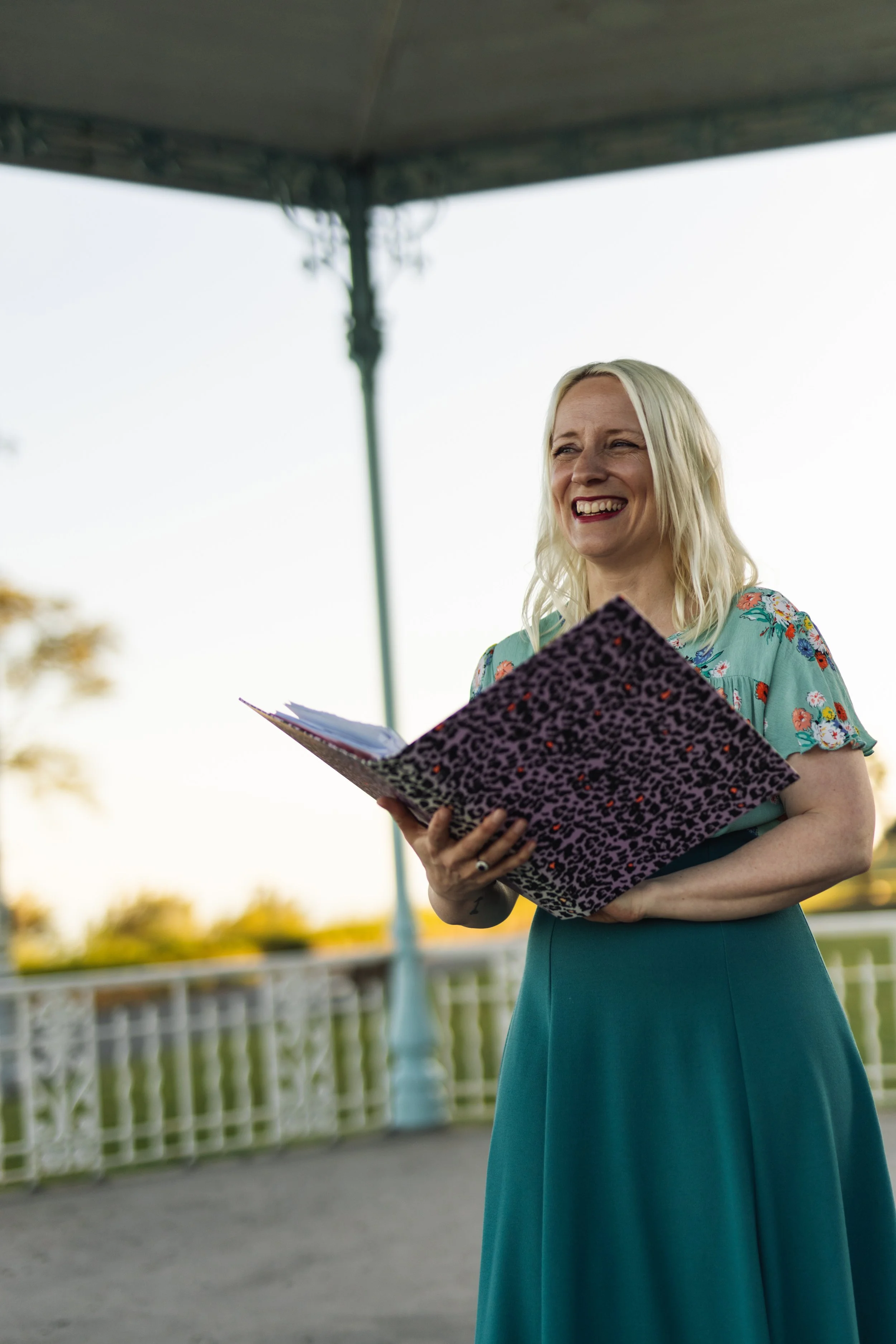 A blonde white woman in a floral blue dress is smiling and holding a purple leopard print document folder