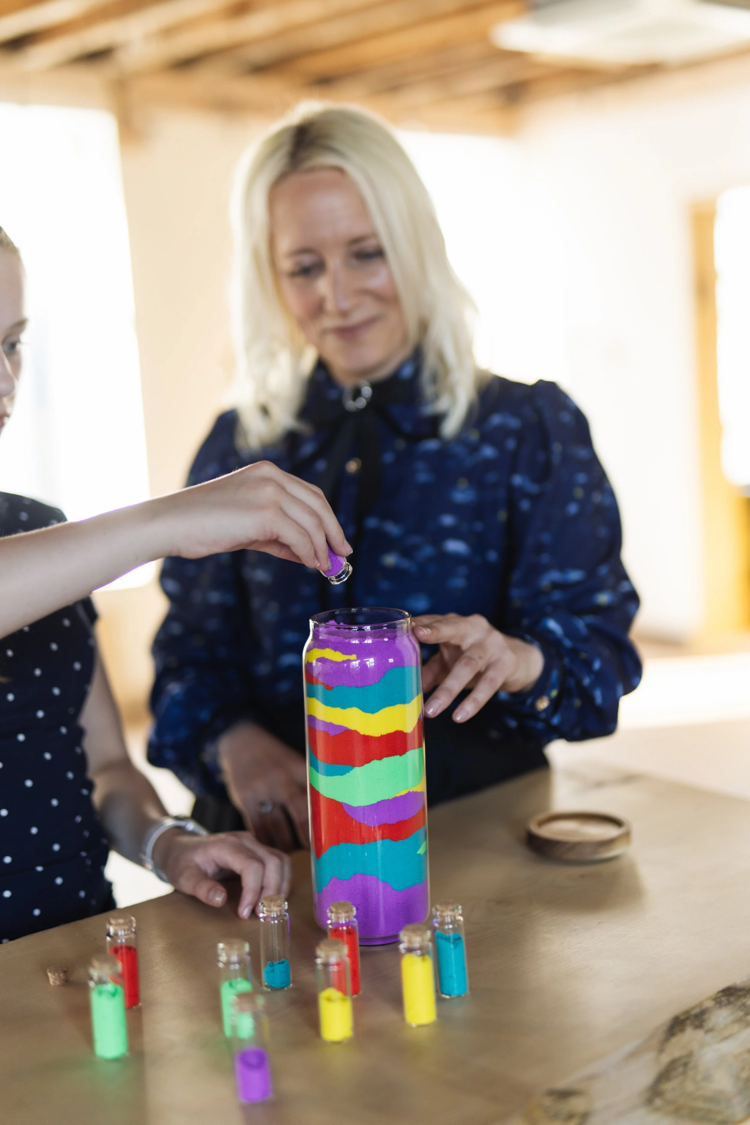 A blonde white woman shows a young girl how to pour purple sand into a large vase with multi-coloured layers of sand in it