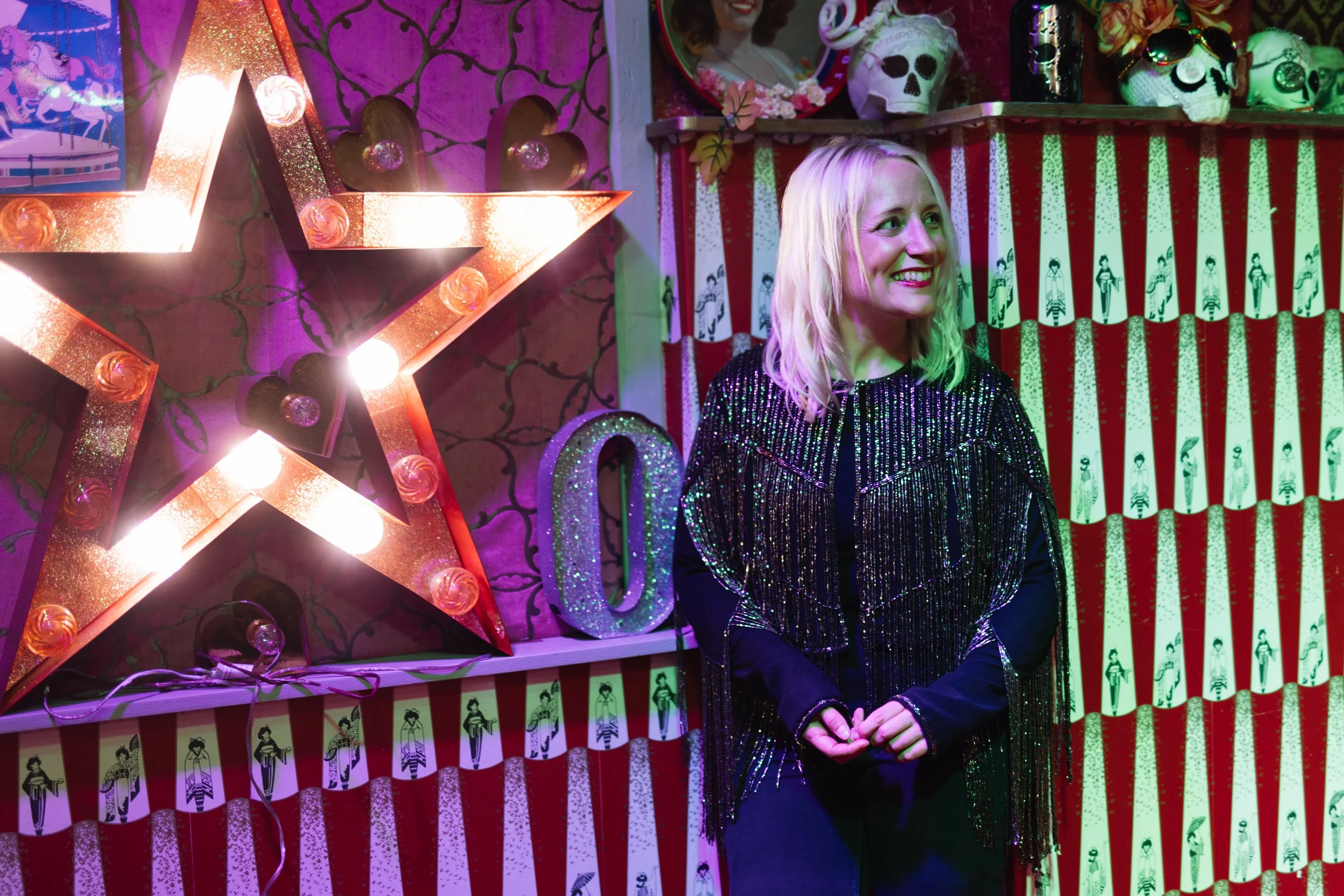 The image shows a blonde haired, white woman wearing a black jacket with sparkly tassels. She is smiling and standing next to a large red metal star with lightbulbs around the outline, and the walls around it are red and white striped like a circus.