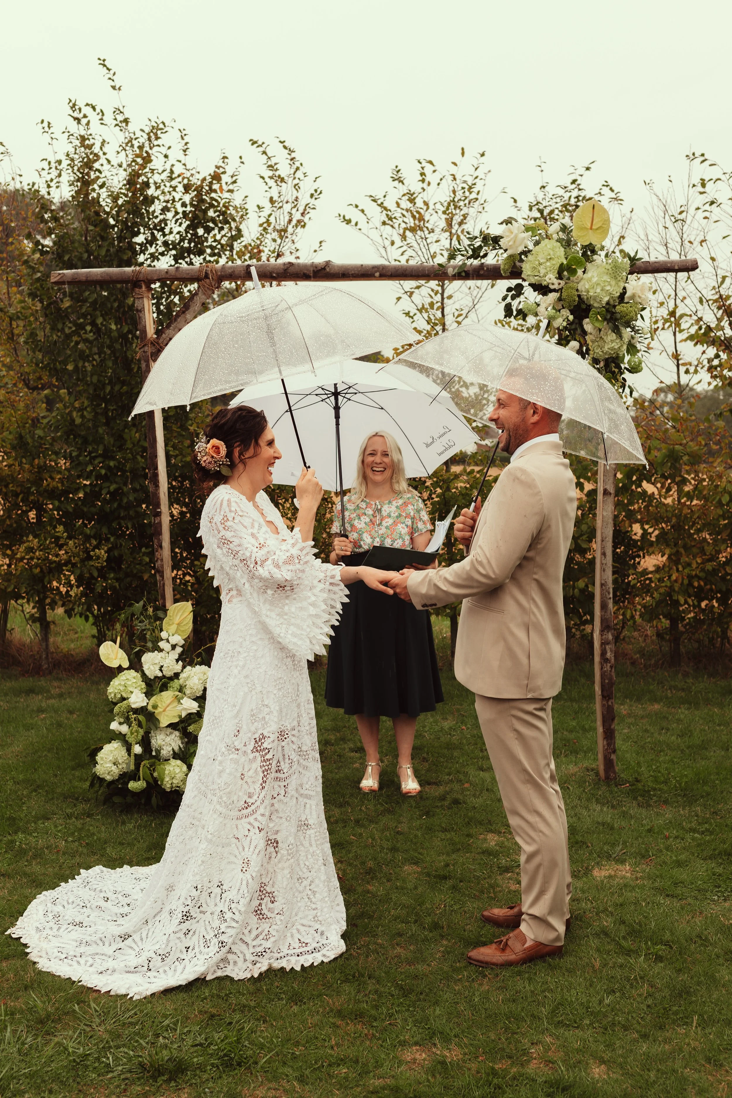 A blonde celebrant laughs with a male/female couple she is marrying. The woman is white with dark hair and a white dress, the man is white and fair haired with a light suit.