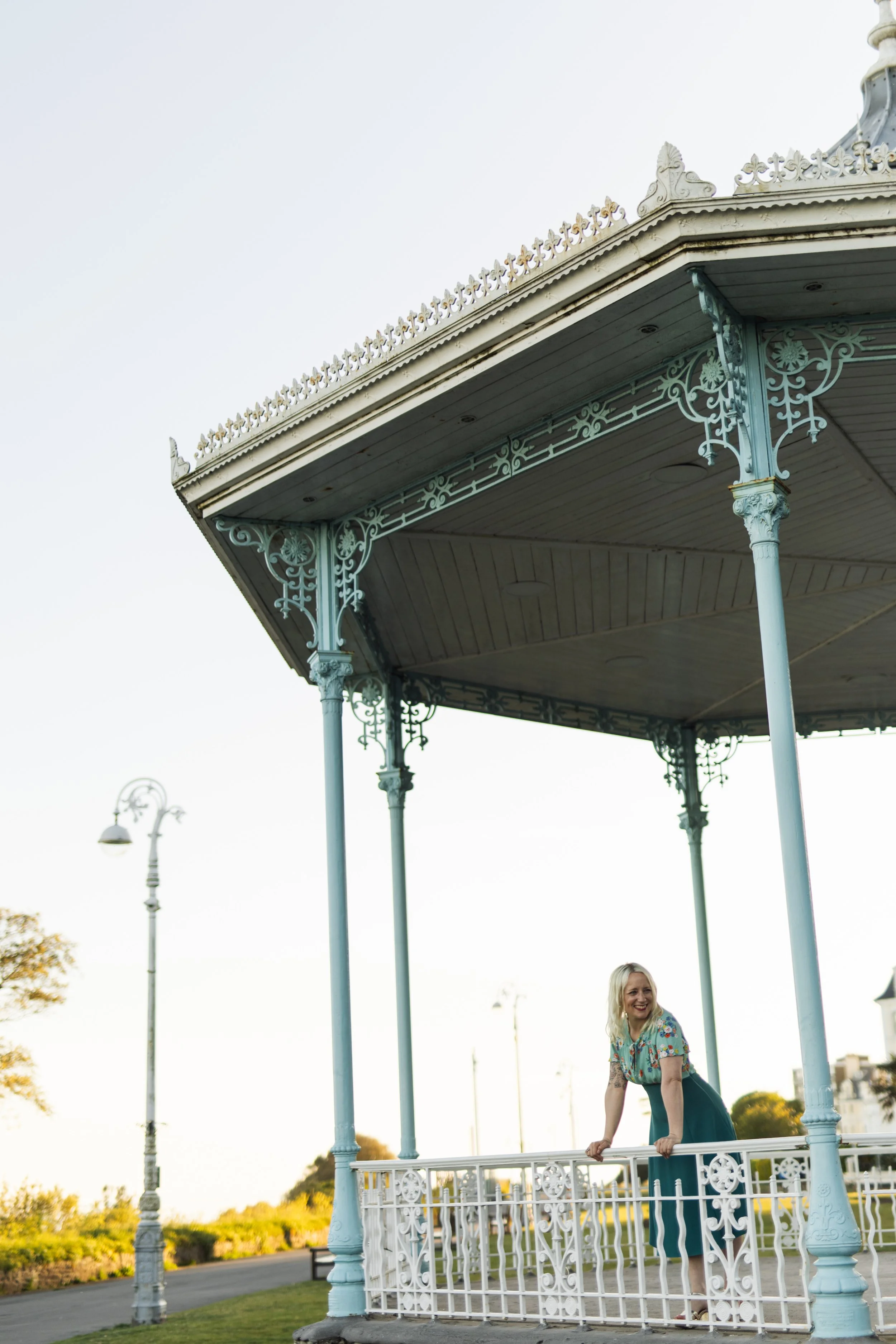 A blonge haired white woman in a blue floral dress leans over the edge of a Victorian bandstand, smiling at something off to the right of the image.