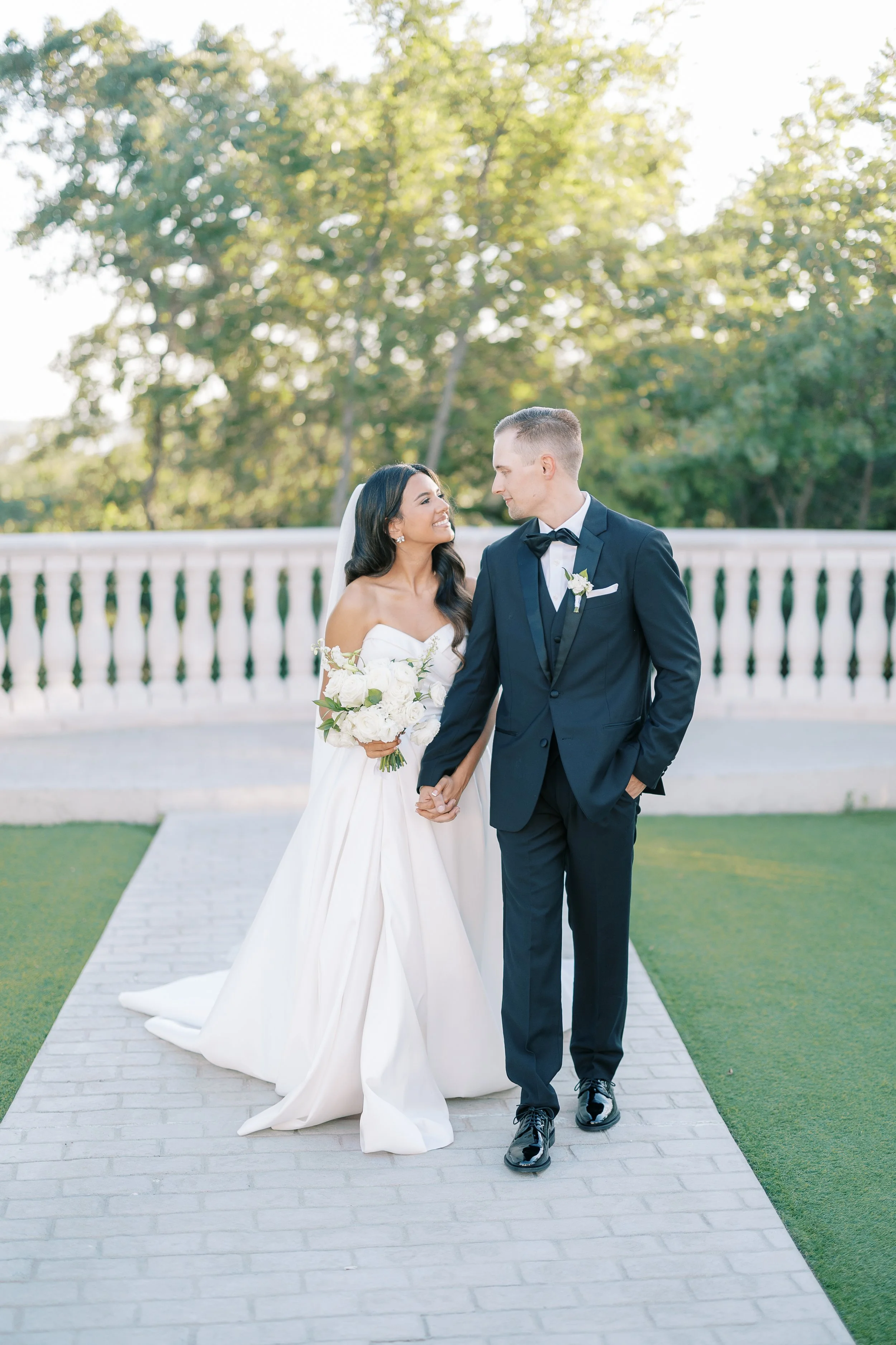A bride and groom holding hands on a stone pathway outdoors, with a white decorative fence and green trees in the background during daylight.