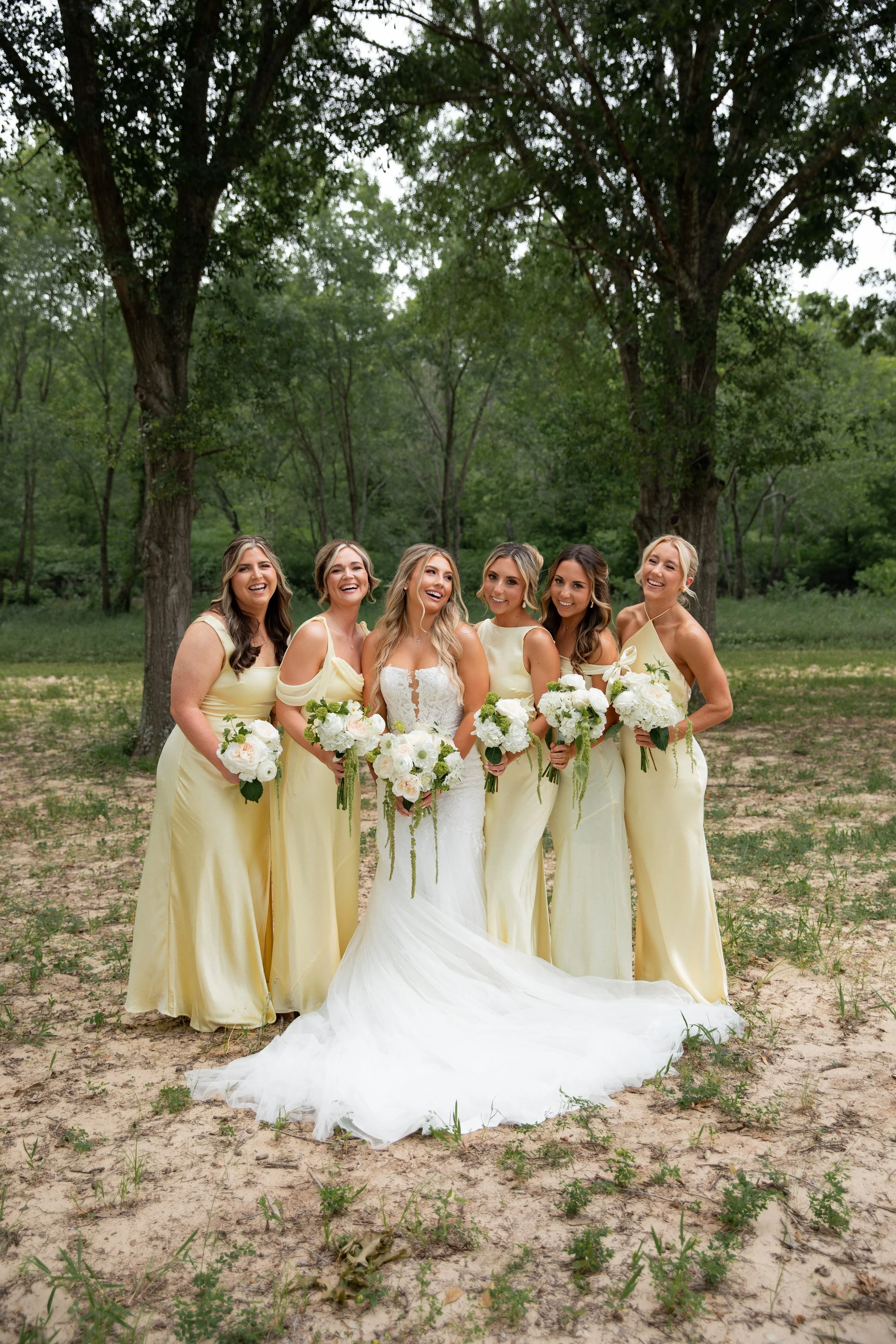 A group of six women in yellow dresses, with one in a white wedding gown, standing outdoors on a grassy area with trees in the background, smiling and holding bouquets.