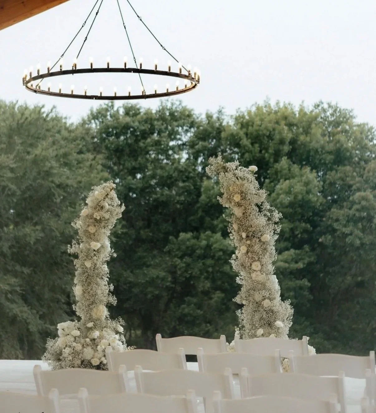 Indoor wedding ceremony setup with floral arches, empty chairs, and a large circular chandelier hanging from the ceiling, with trees visible outside through large windows.