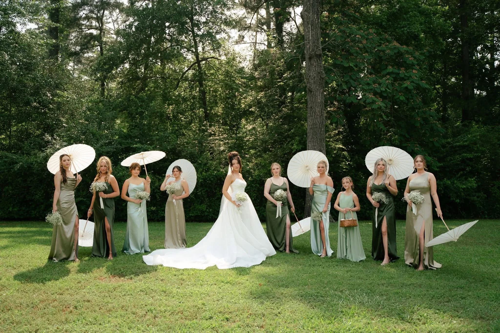A group of women in formal dresses and holding umbrellas and bouquets standing outdoors in a lush green park with tall trees.