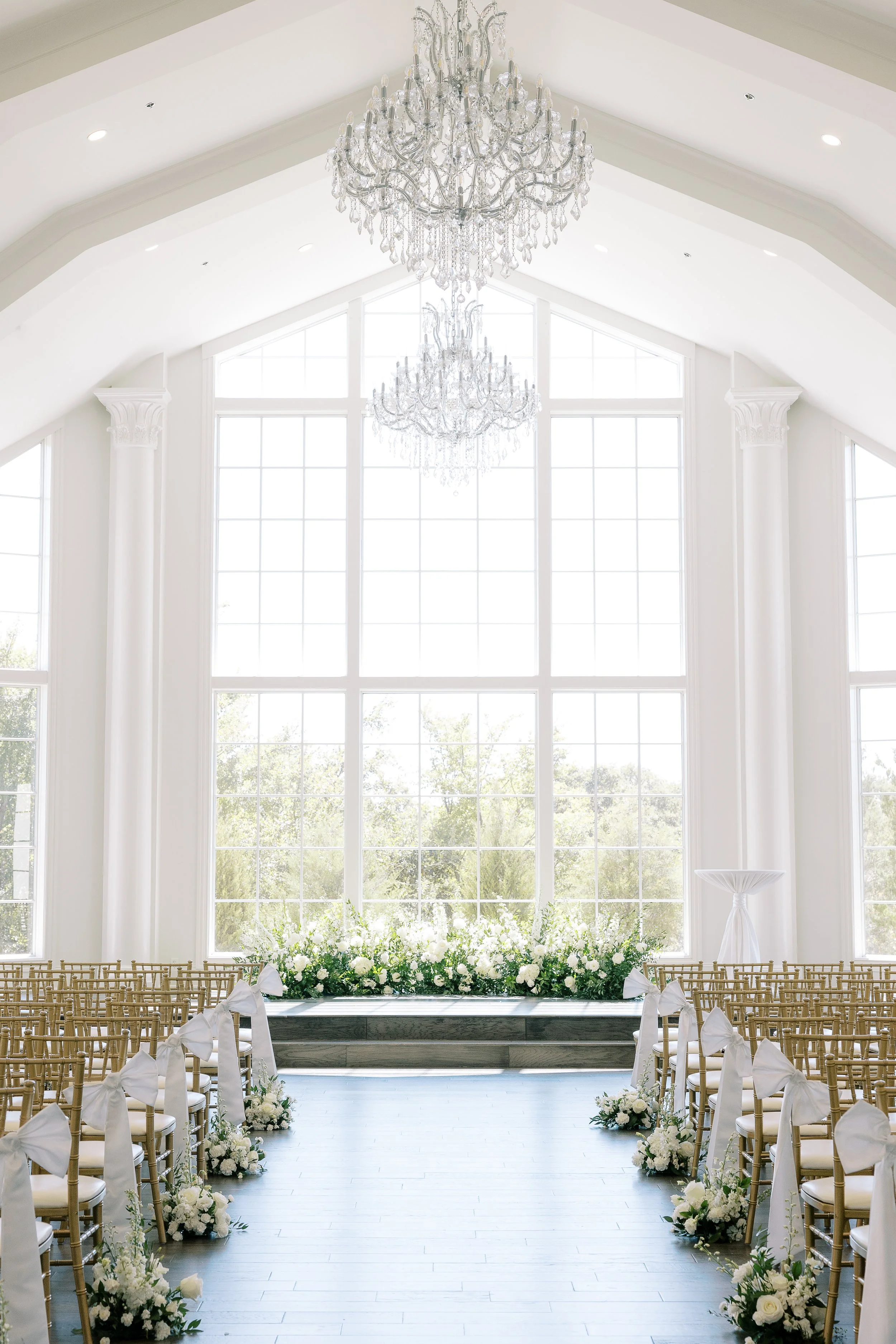 A wedding ceremony setup in a bright, elegant room with large windows, chandeliers, rows of chairs with white bows, and a floral aisle leading to an altar with white flowers.