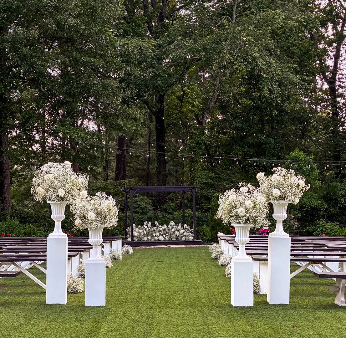 Outdoor wedding ceremony setup with white floral arrangements on pedestals, wooden benches, a grassy aisle, and a wedding arch surrounded by trees and string lights.