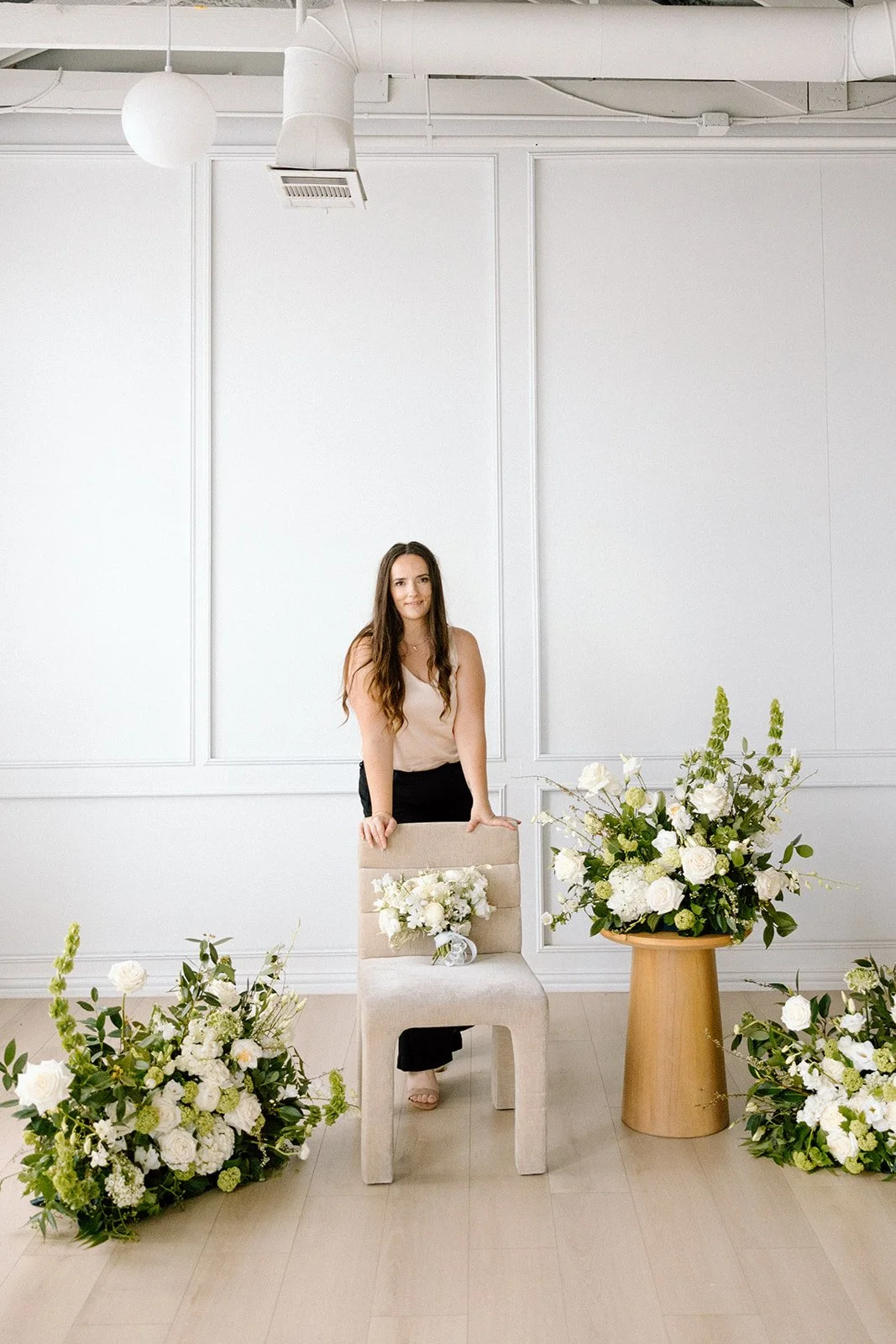 A woman standing behind a beige chair with a floral arrangement on it, in a room with white walls and large floral displays on the floor and in a large wooden vase.
