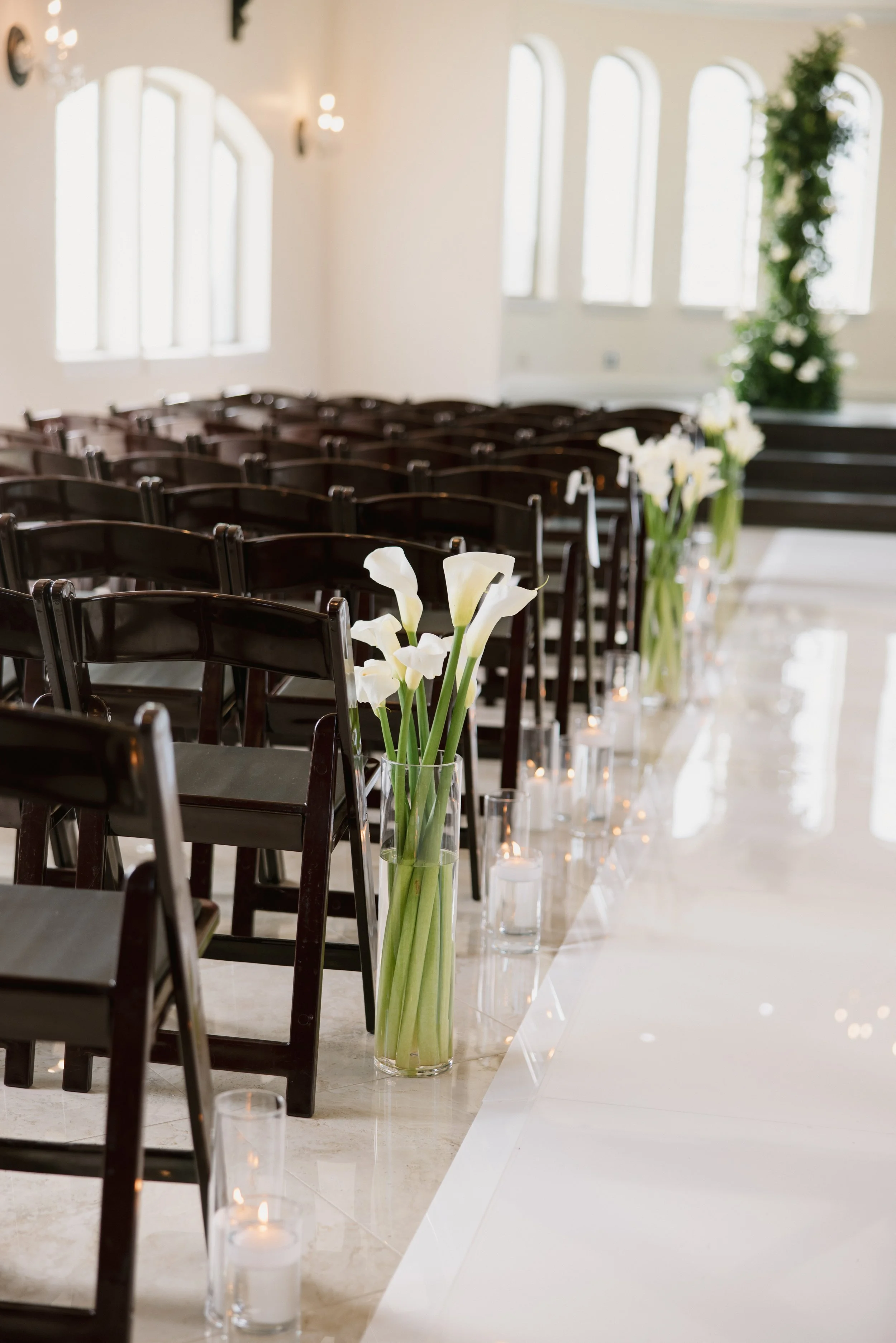 Indoor wedding or event setup with a white aisle, large windows, white flowers in a tall clear vase, and tealight candles in glass cylinders along the aisle.