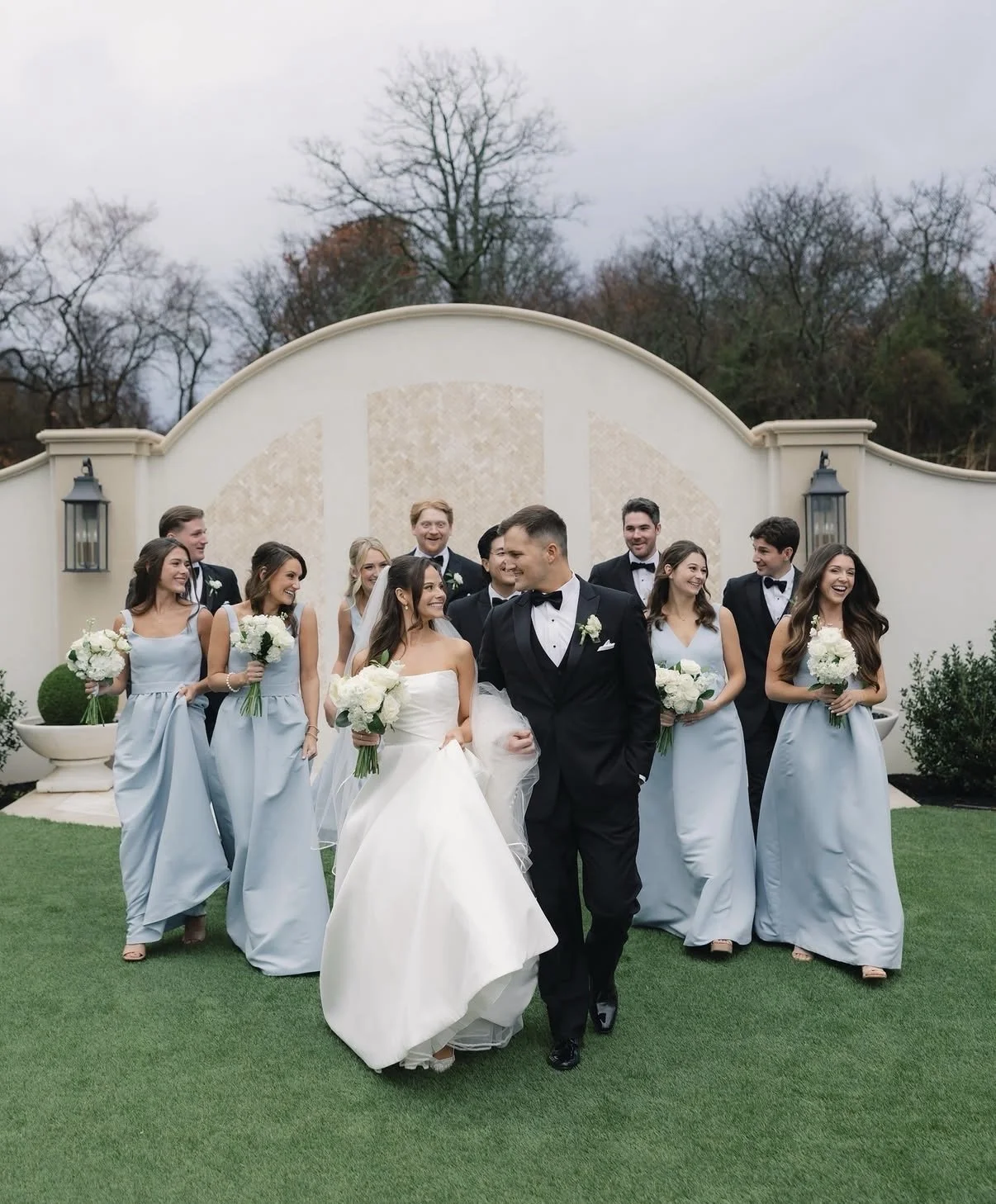 A wedding party, including the bride in a white gown and the groom in a black tuxedo, walking outdoors on a well-manicured lawn. Bridesmaids in light blue dresses and groomsmen in black tuxedos follow behind, all holding white bouquets and smiling.