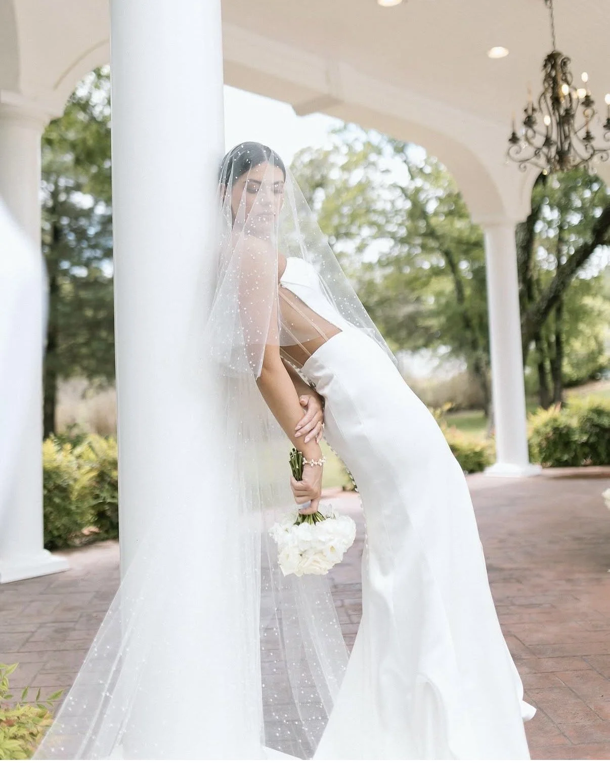 A bride in a white wedding dress holding a bouquet of white flowers, standing outdoors near white columns with greenery and trees in the background.