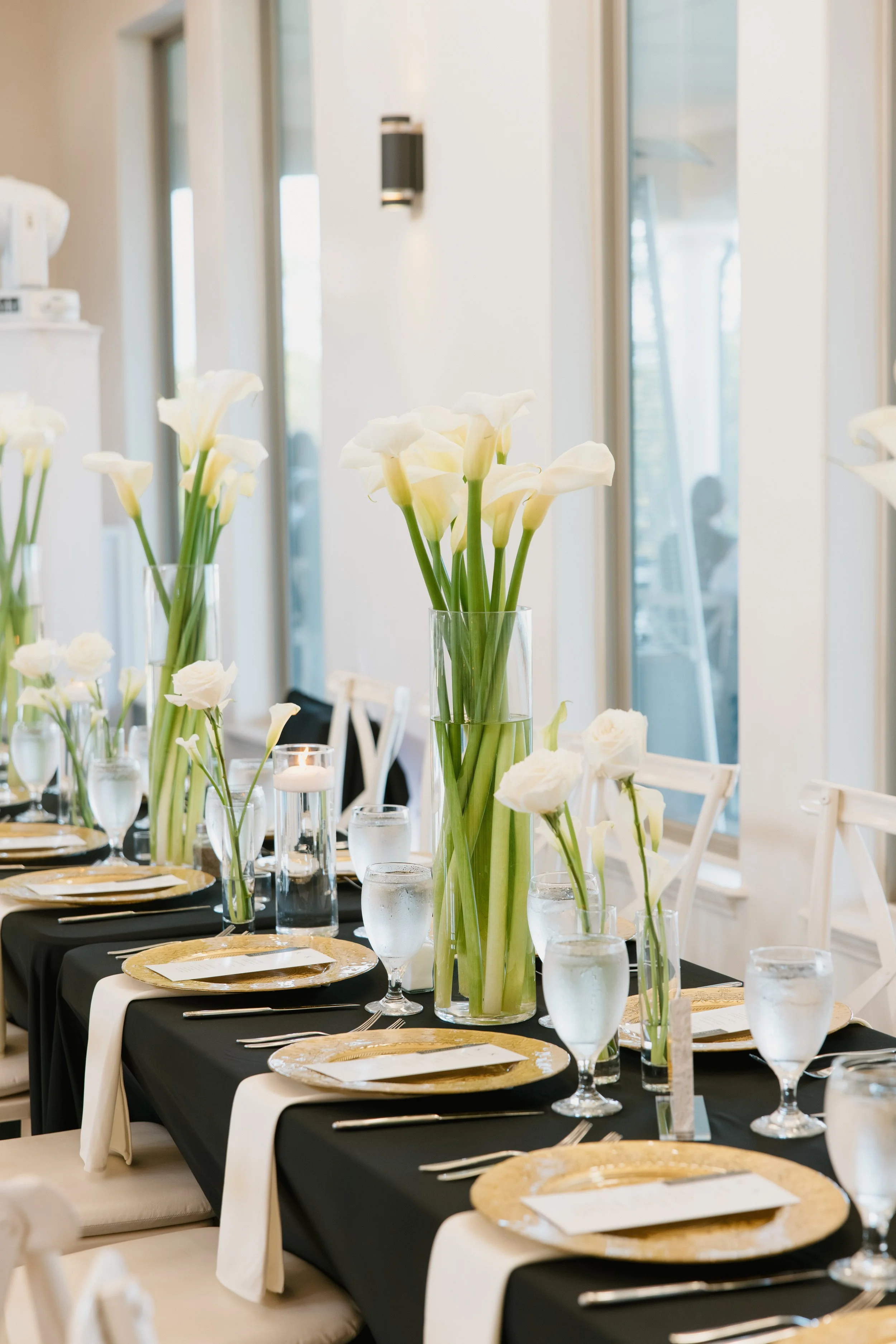 Elegant dining table decorated with tall white calla lilies in vases, set with gold chargers, white plates, silverware, and water glasses, in a bright room with large windows.