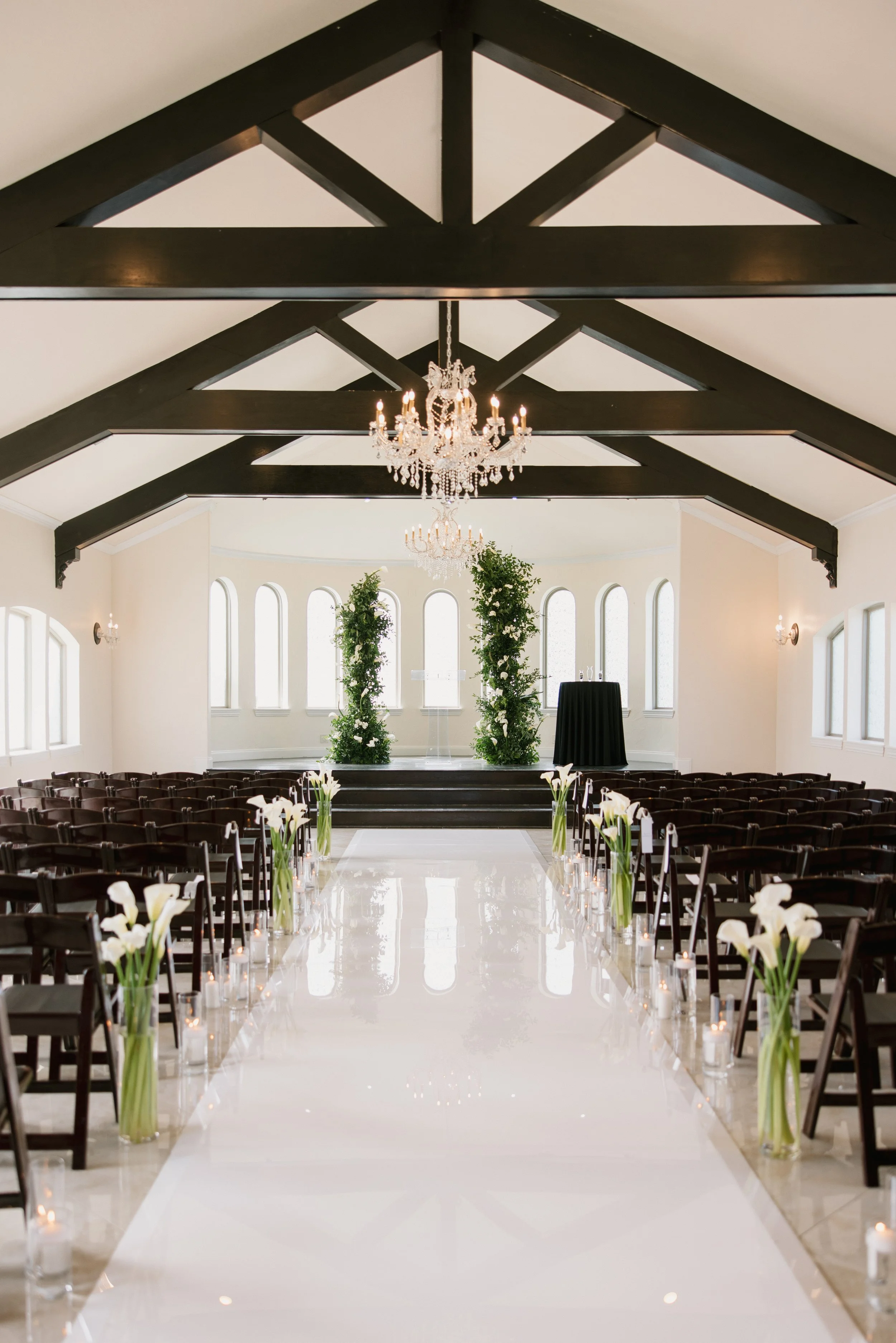 An elegant wedding aisle with white flowers in tall vases on either side, lit candles in glass holders, and a shiny white flooring leading to an altar with two large green plants and chandeliers hanging from a ceiling with dark wooden beams.
