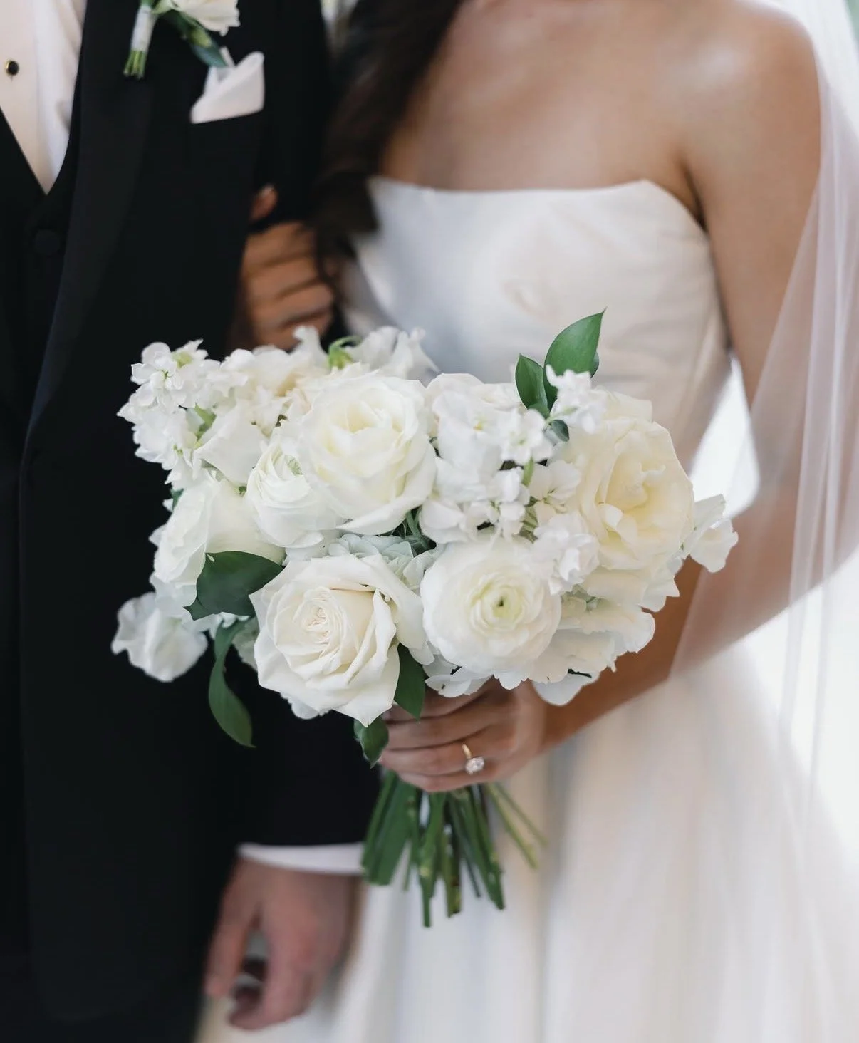 Bride holding a bouquet of white roses and flowers, standing next to groom in a black tuxedo.