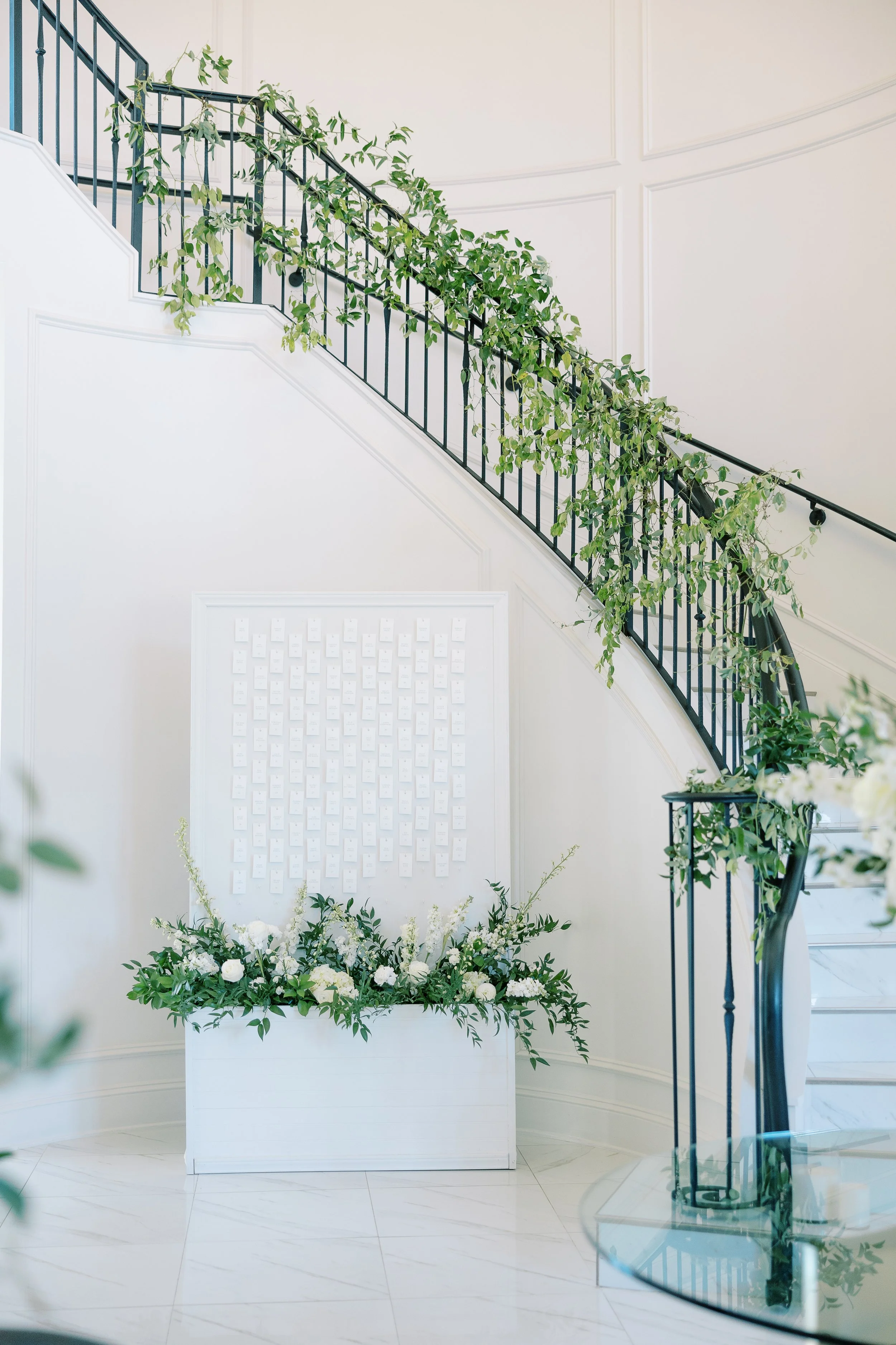 Elegant indoor space with a staircase railing decorated with green leafy foliage, a white wall with a framed display of small mostly blank cards, and a floral arrangement of white flowers and greenery on a white stand.