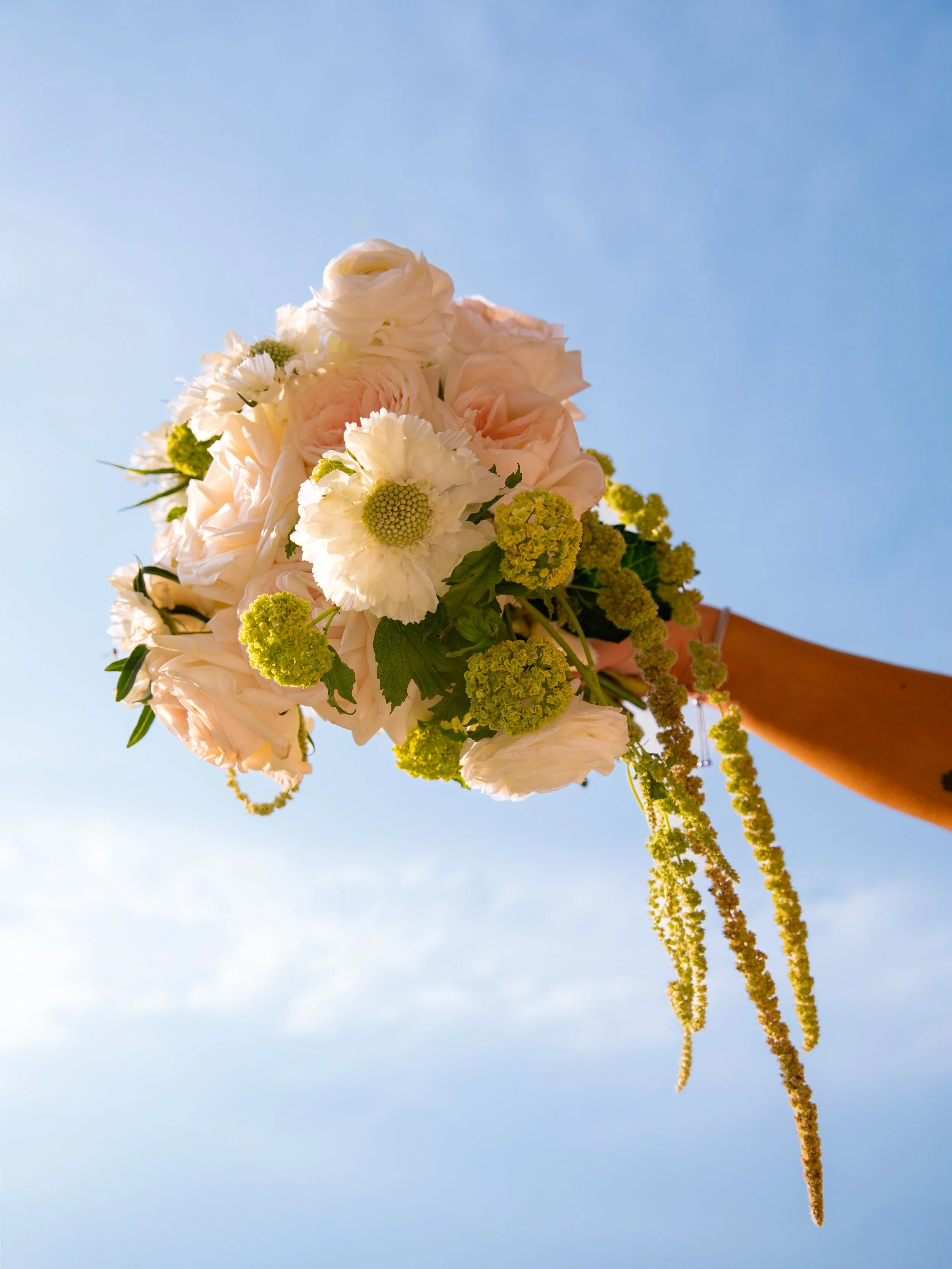 A bouquet of flowers held against a clear blue sky, featuring white roses, light pink roses, white daisies, and yellow-green filler flowers.