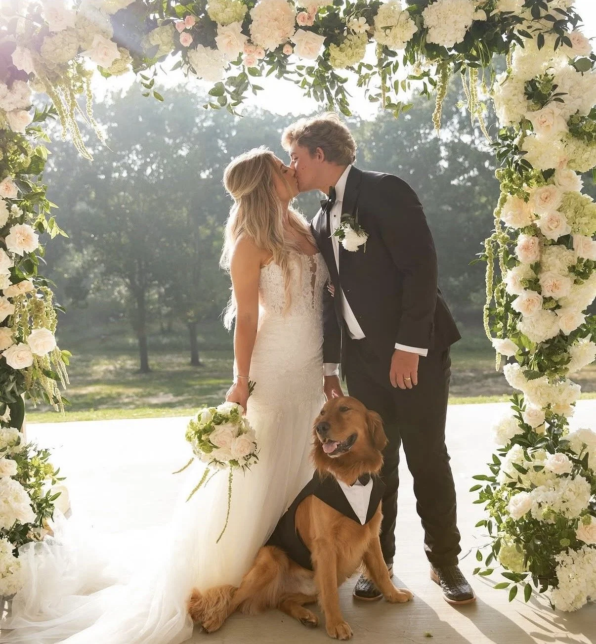A bride and groom sharing a kiss during their outdoor wedding ceremony, standing under a floral arch with white and pale pink flowers, with a golden retriever wearing a tuxedo sitting at their feet.