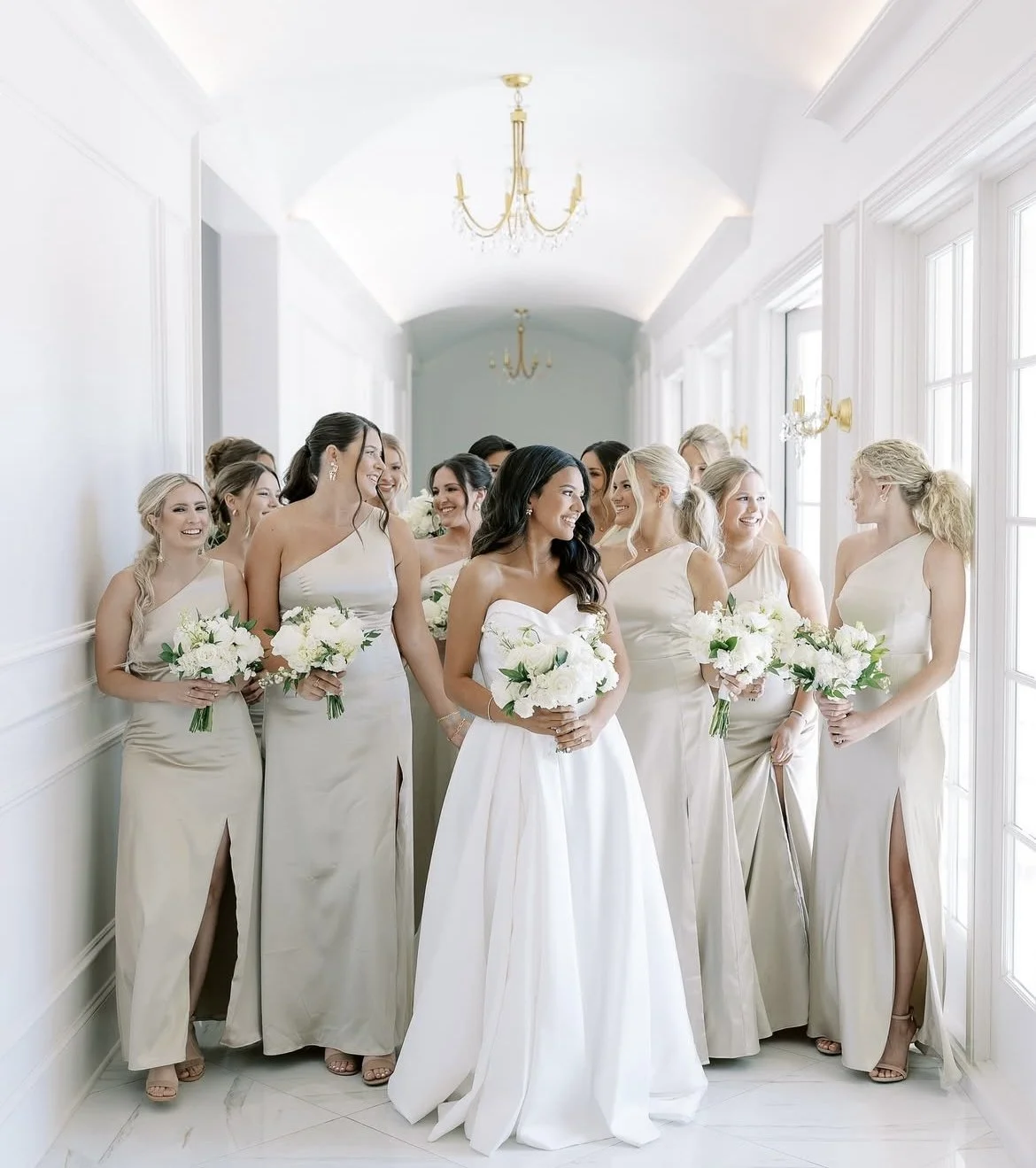 A group of women, likely bridesmaids and a bride, standing in a bright, elegant hallway with large windows, white walls, and chandeliers, all holding white flower bouquets and dressed in cream-colored dresses.