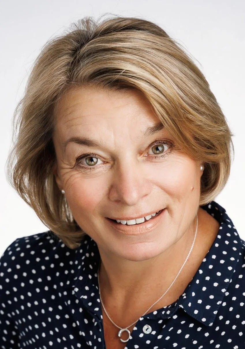 Close-up of a middle-aged woman with blonde hair, smiling, wearing pearl earrings, a silver necklace, and a navy blue shirt with white polka dots, against a plain white background.