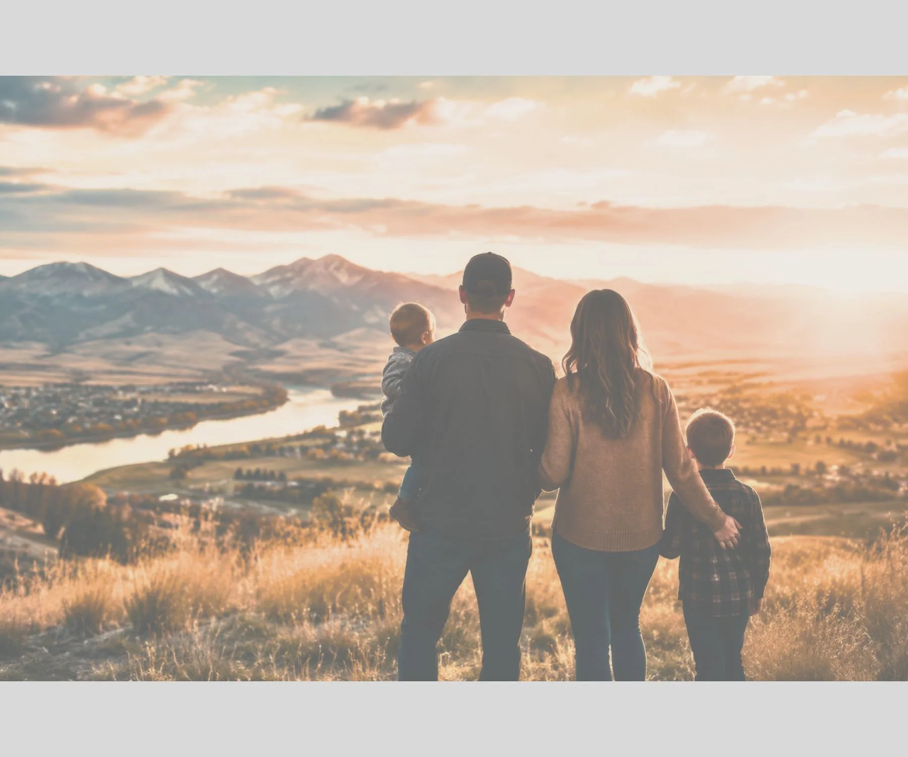 A family of four holding hands and walking together outdoors during sunset, with mountains, a river, and a cloudy sky in the background.