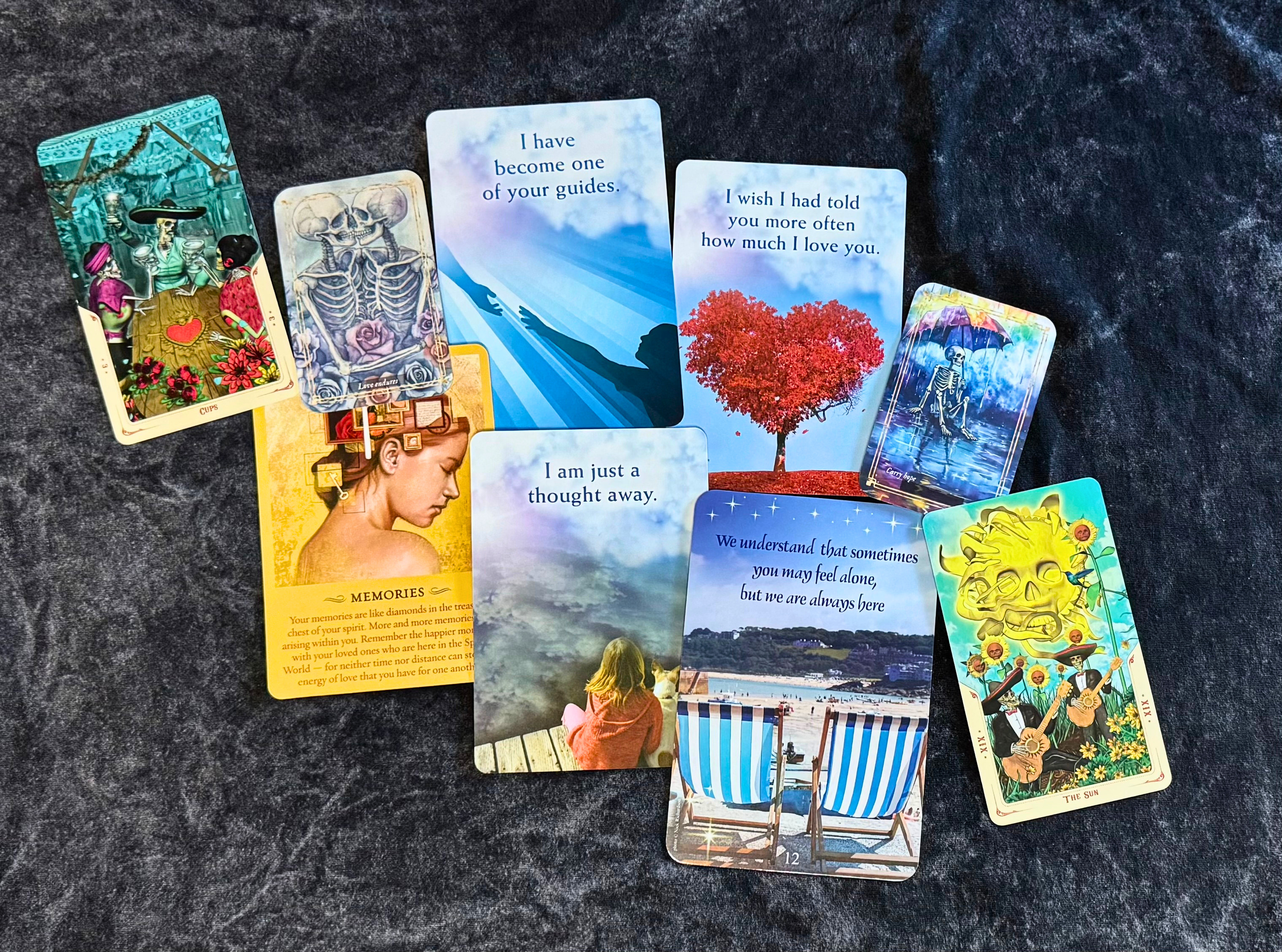 A collection of tarot and mediumship cards laid out on a dark surface, featuring images of skeletons, a girl sitting on a bench, a rainbow skull, and a colorful skeleton playing guitar, with some cards displaying messages and symbols.