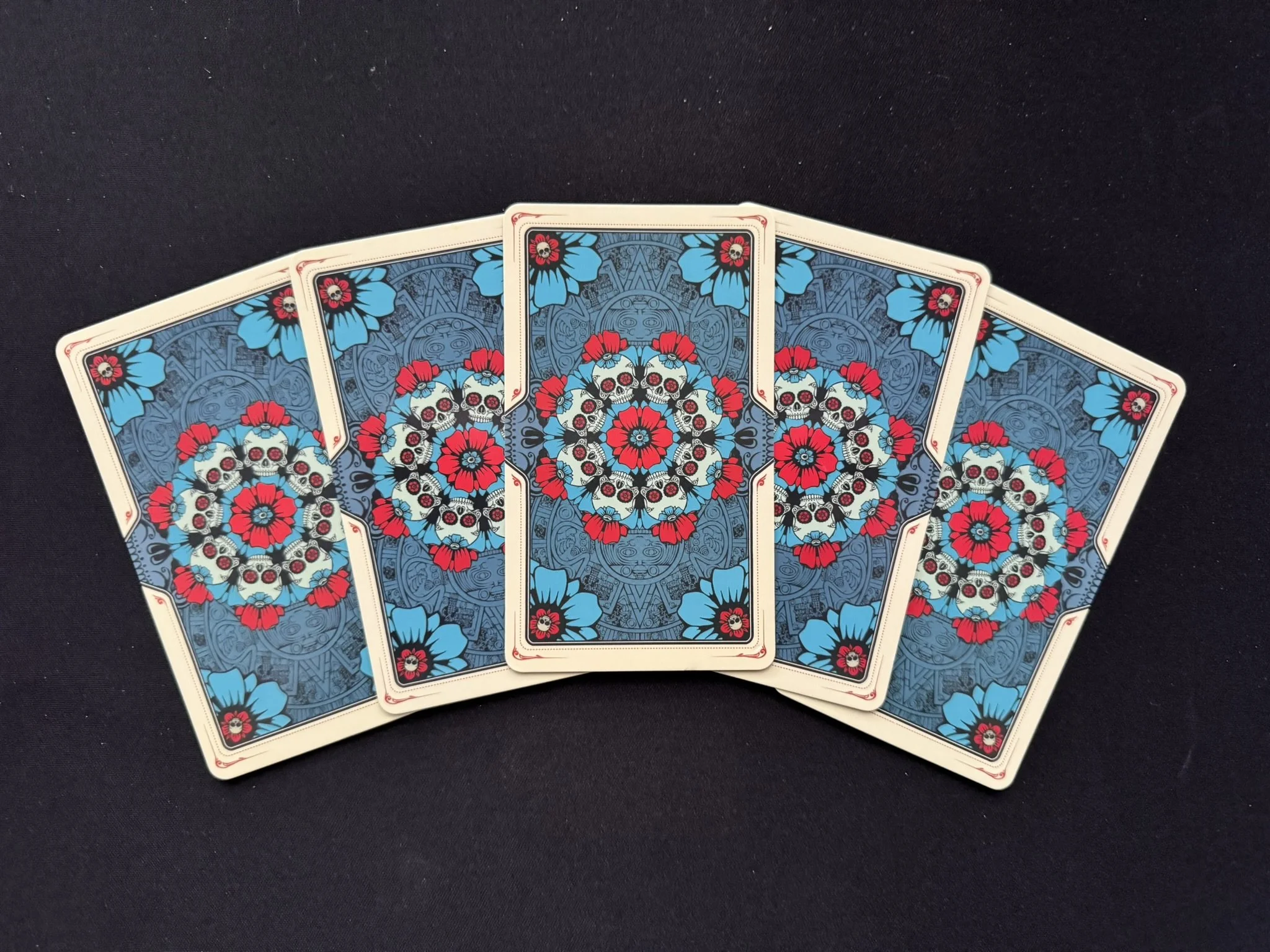 Seven tarot or playing cards with a symmetrical, colorful skull and floral design on their backs, fanned out on a black surface.